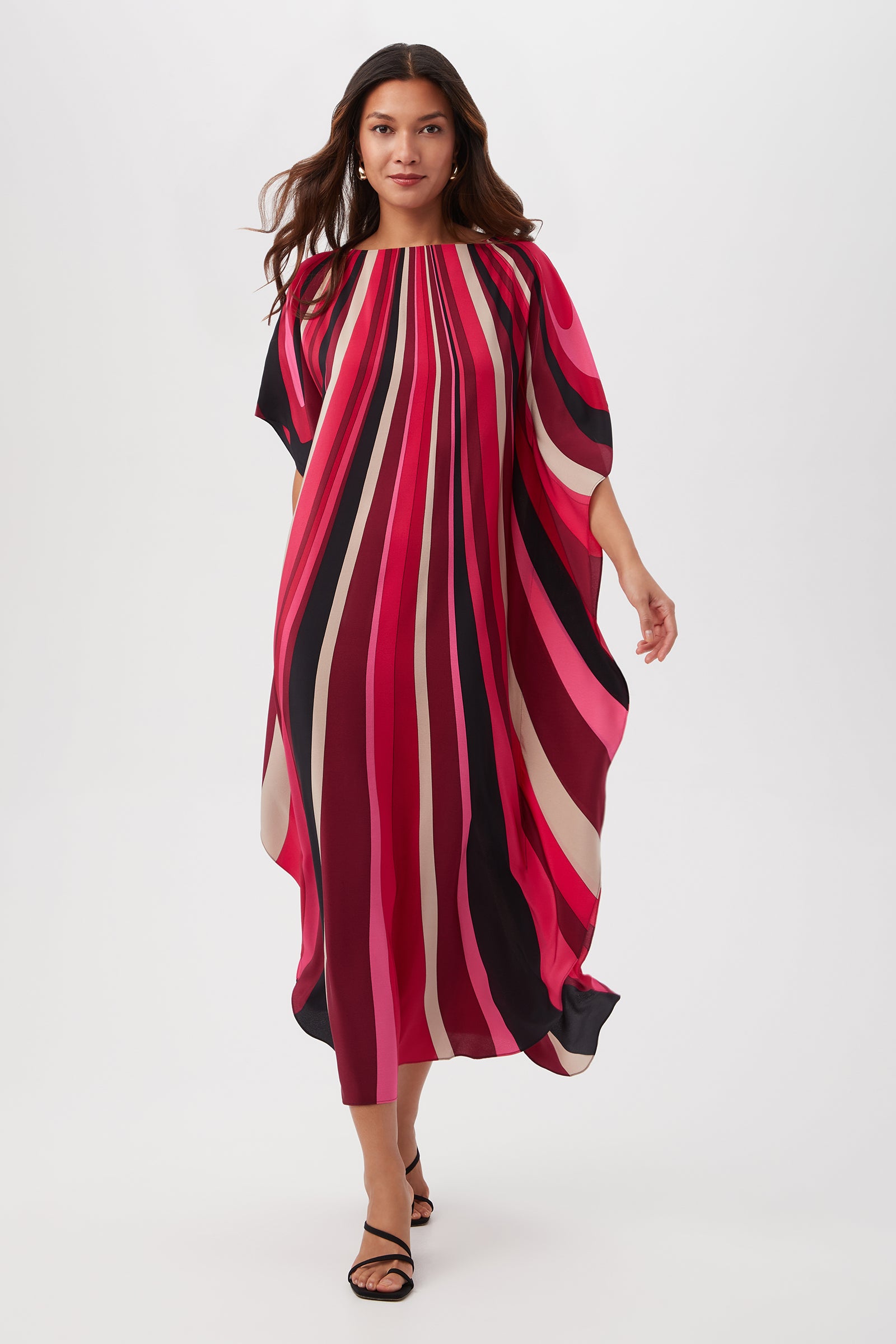 A woman walks confidently, her hair in loose waves, wearing the Jalani Maxi Caftan Dress - Wynwood Waves Silk in Fairy Fuchsia Multi—its bold, flowing stripes of red, pink, black, and white making a vibrant statement.