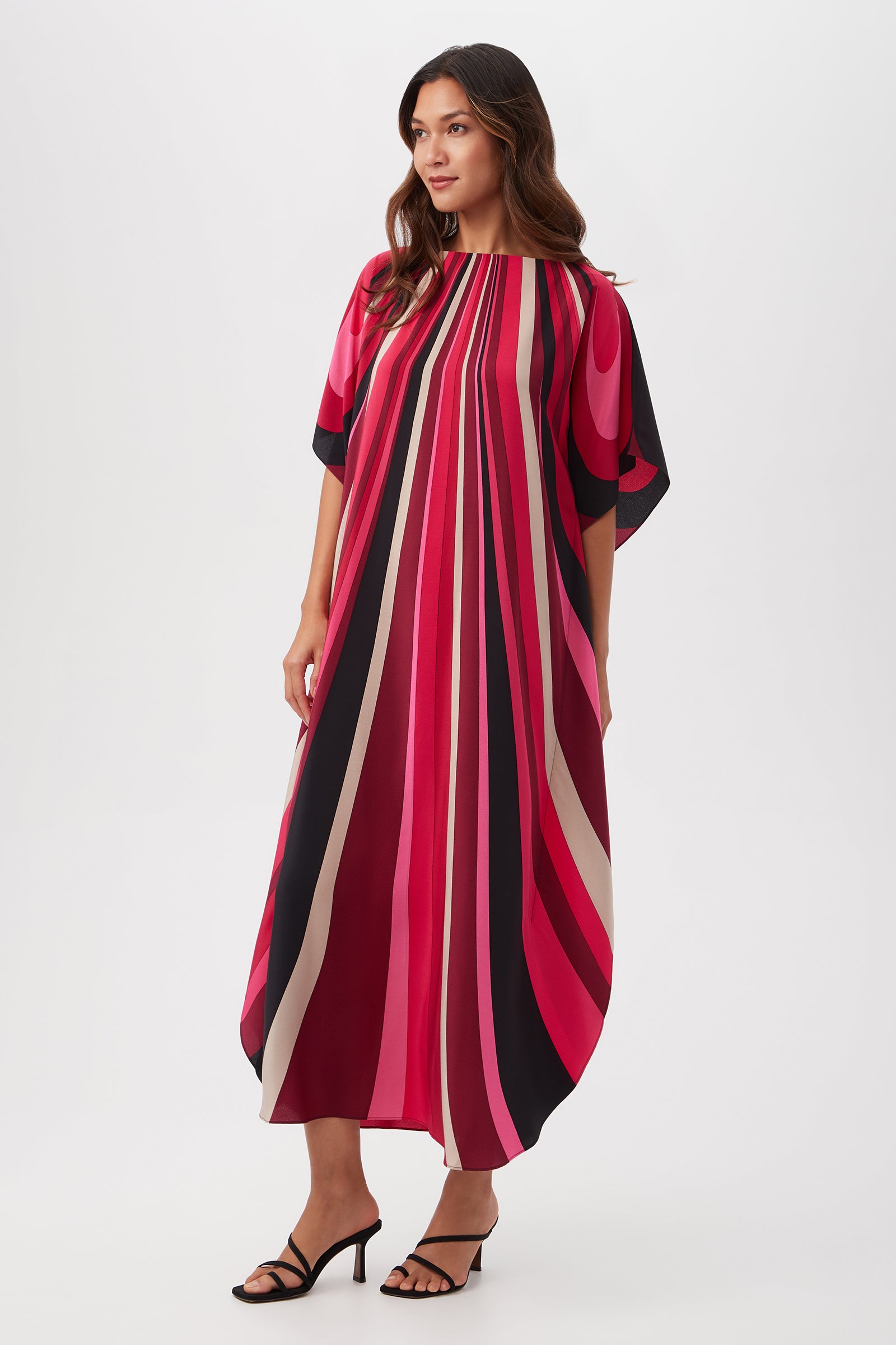 A woman wears the Jalani Maxi Caftan Dress - Wynwood Waves Silk, a loose, ankle-length dress with bold vertical pink, red, black, and beige stripes. It features wide short sleeves and a round neckline. She pairs it with black heeled sandals against a white background.