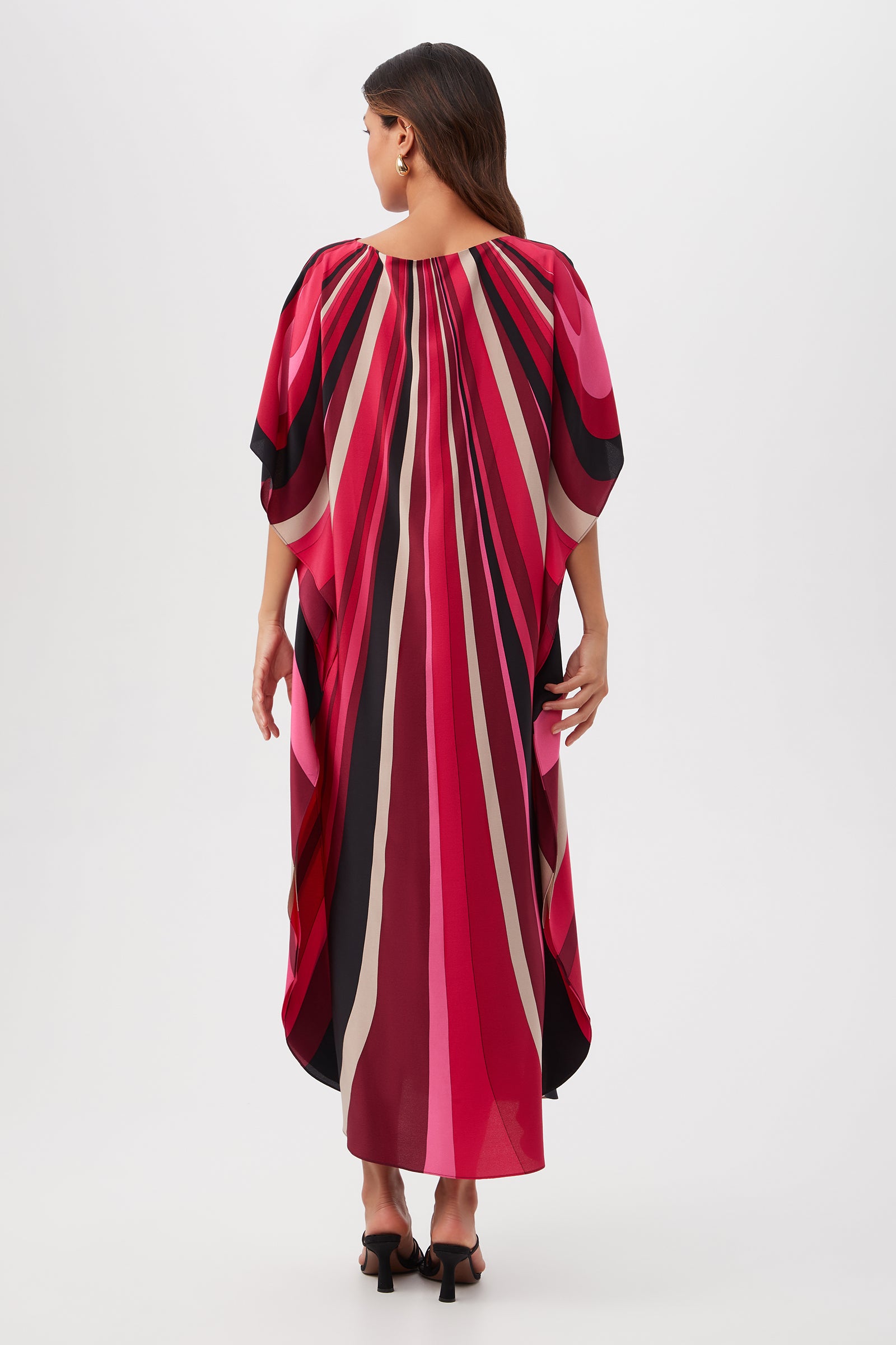 A woman with long, dark hair stands facing away, wearing the Jalani Maxi Caftan Dress - Wynwood Waves Silk, a loose, flowing dress featuring bold vertical stripes in pink, red, black, and beige. She pairs it with heeled sandals.
