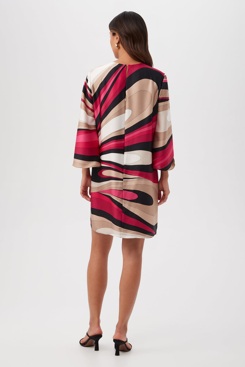 A woman with long hair wears the Nallia Dress - Wynwood Waves Satin Georgette, a loose, knee-length dress with long sleeves and a bold abstract pattern in red, pink, black, and beige tones. She pairs it with black heeled sandals.