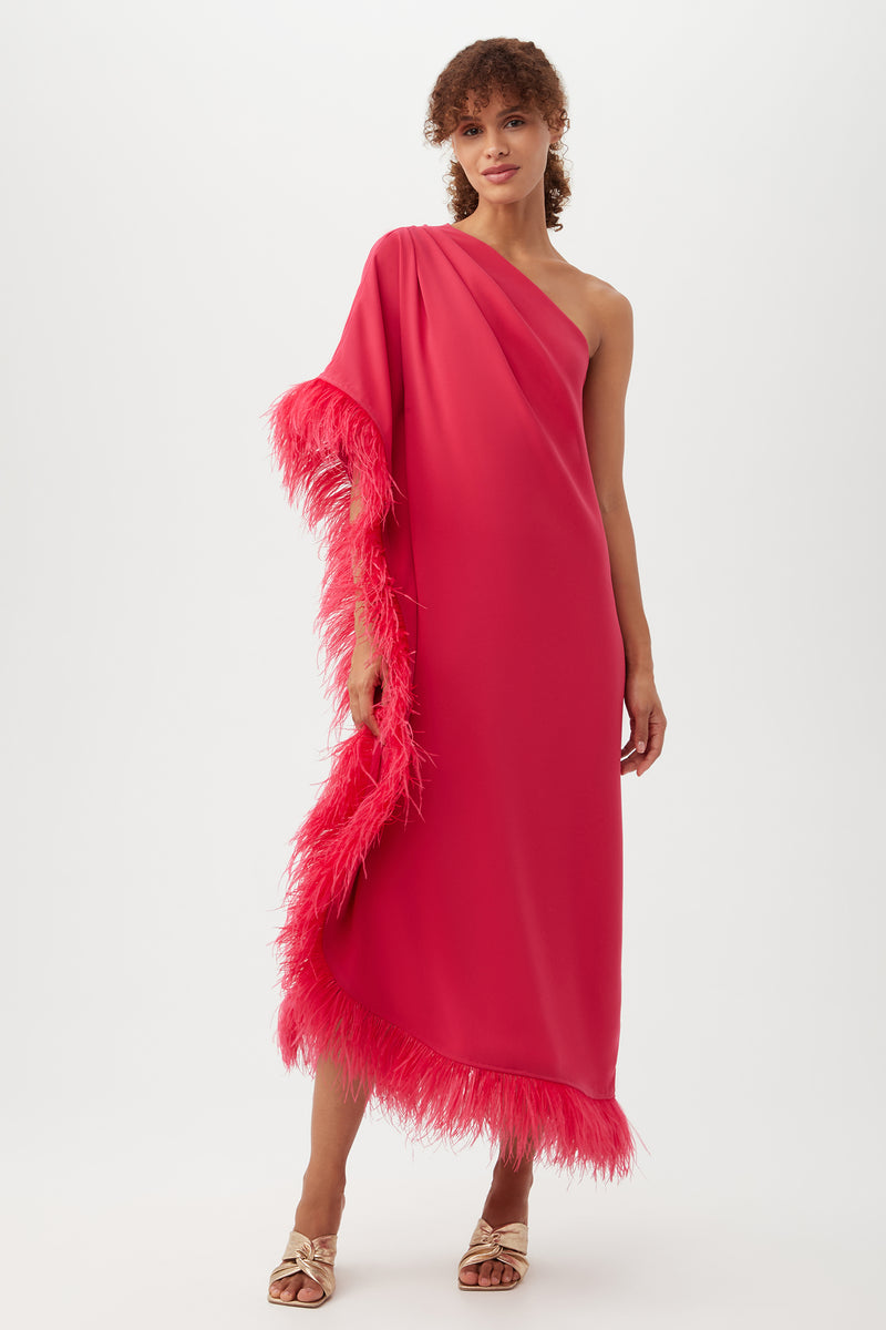 A woman wears the Largo Caftan Dress in Hibiscus, featuring a one-shoulder design and feather trim along the hem and side. She stands against a white backdrop, pairing the dress with light open-toe heeled sandals.
