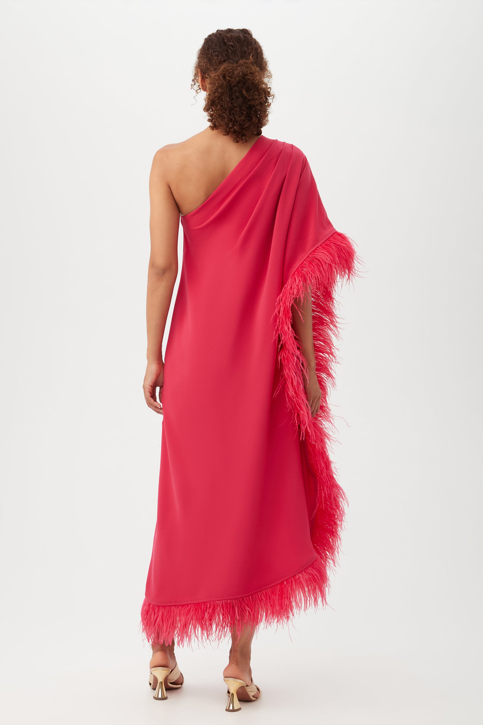 A woman stands with her back to the camera against a plain white background, wearing the Largo Caftan Dress—an ankle-length, one-shoulder bright pink dress featuring feather trim along the edge.