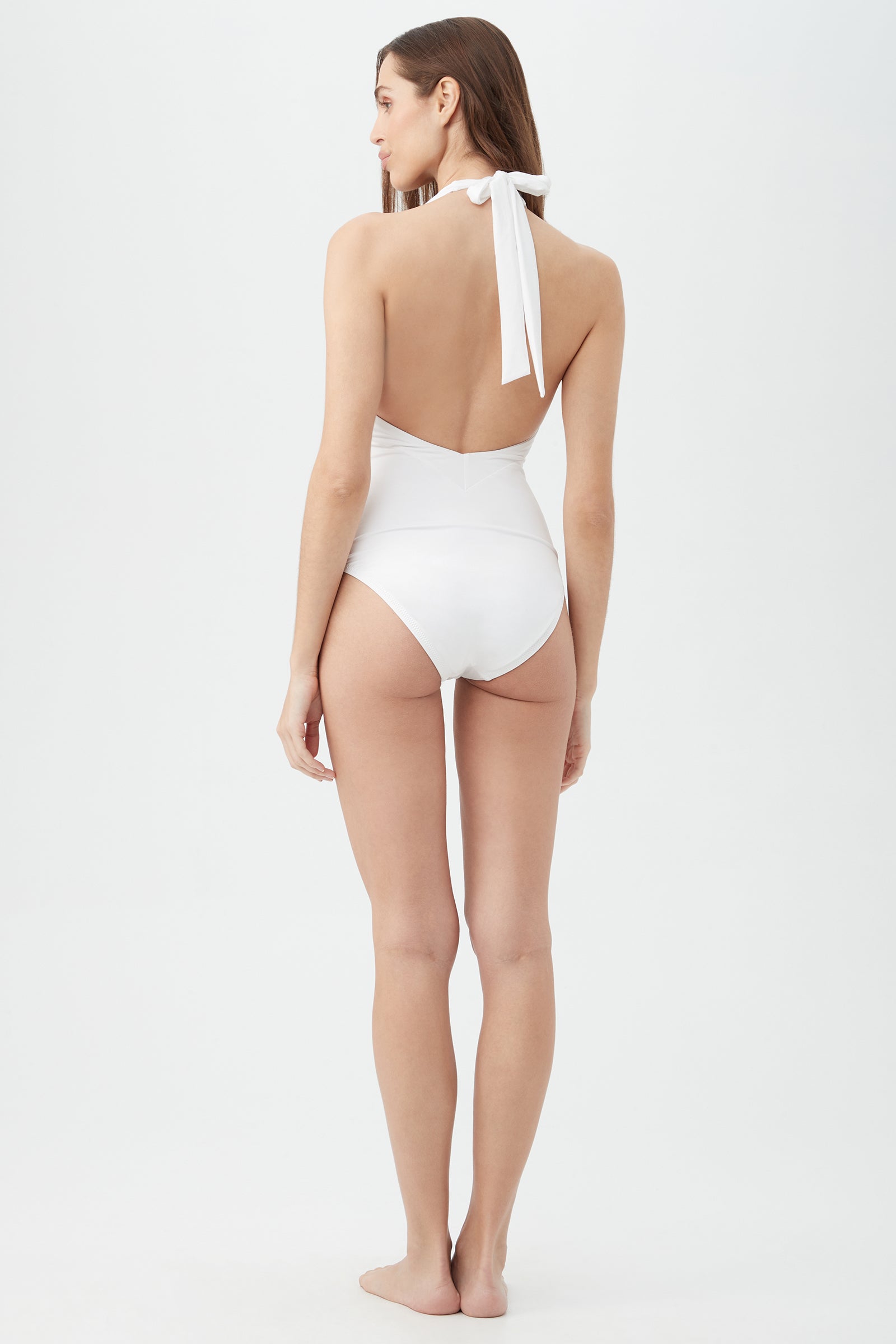 A woman stands barefoot on a white background, facing away, wearing the Women's Monaco Halter Plunge One Piece Swimsuit in white with a neck bow and her straight brown hair down her back.