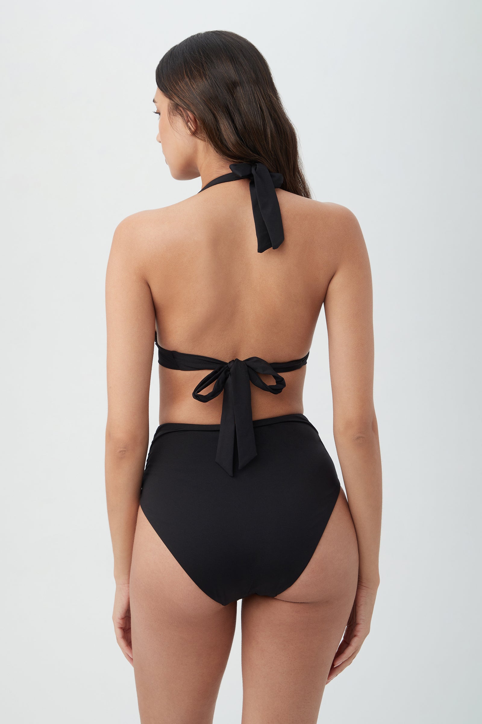 A woman with long dark hair is seen from behind, wearing the Women's Monaco Ring Halter Swim Top in black with high-waisted bottoms and tied bows at the neck and back, standing against a plain light background.