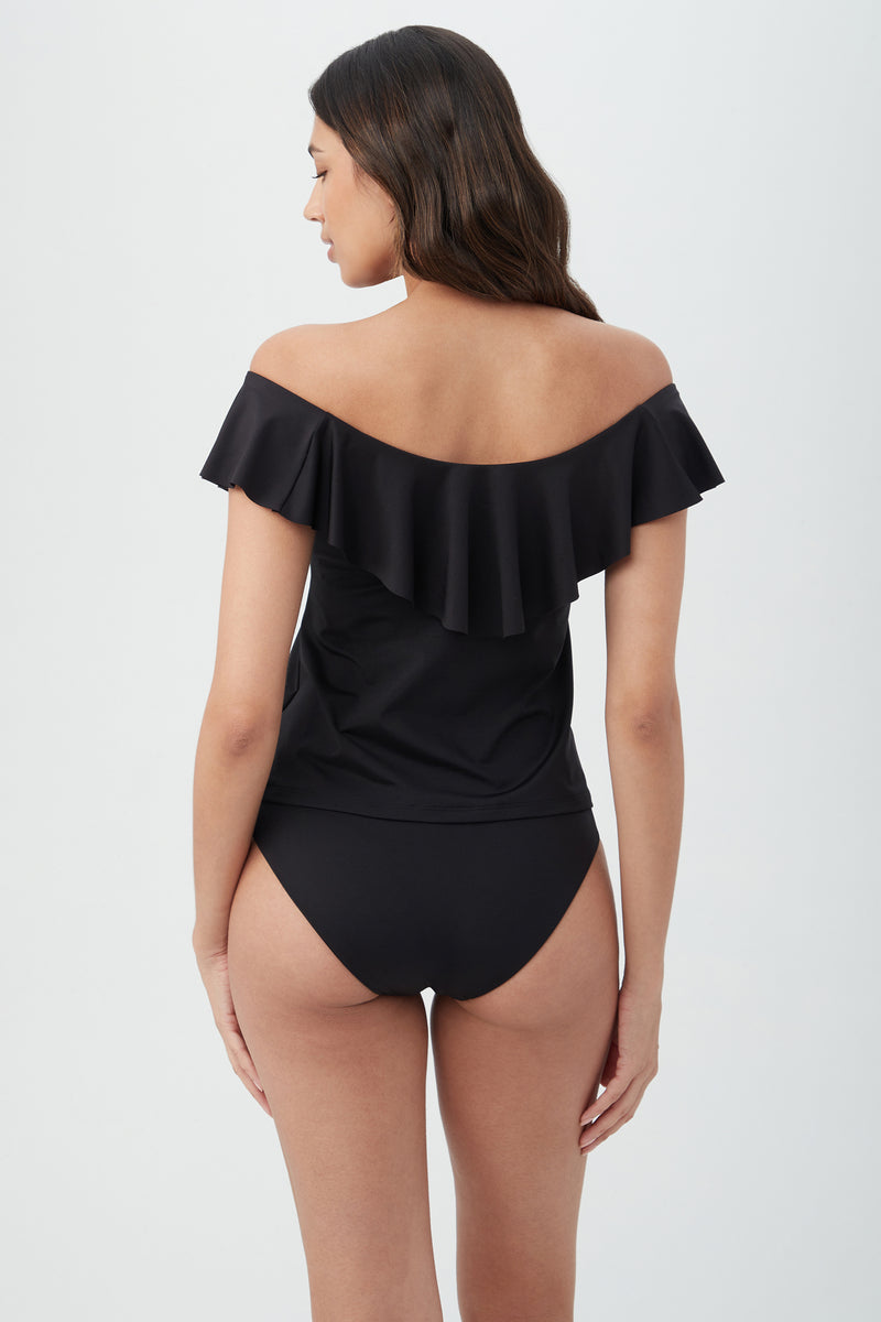 A woman with long, wavy brown hair stands with her back to the camera against a light background, wearing the Women's Monaco Off The Shoulder Ruffle Tankini Swim Top in black.