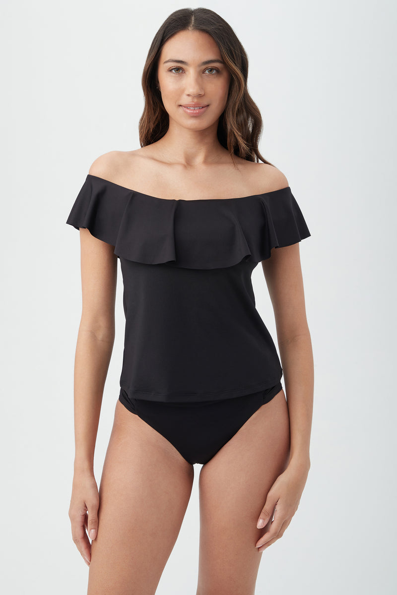 A woman with long, wavy brown hair wears the black Women's Monaco Off The Shoulder Ruffle Tankini Swim Top and matching bikini bottoms, standing against a plain light background.
