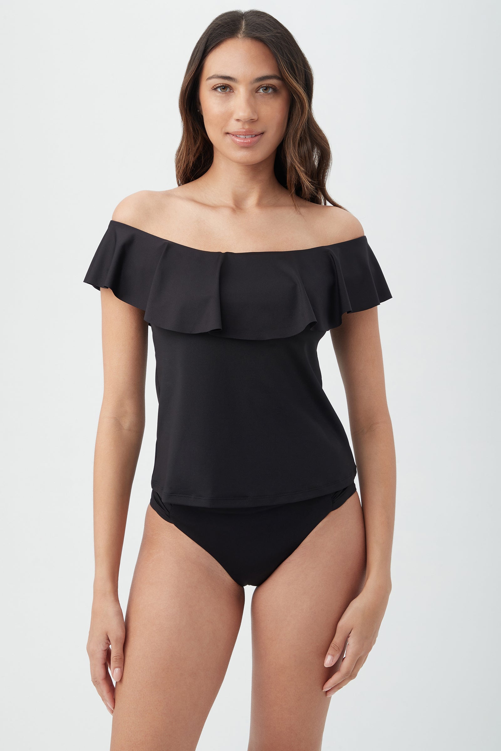 A woman with long, wavy brown hair wears the black Women's Monaco Off The Shoulder Ruffle Tankini Swim Top and matching bikini bottoms, standing against a plain light background.