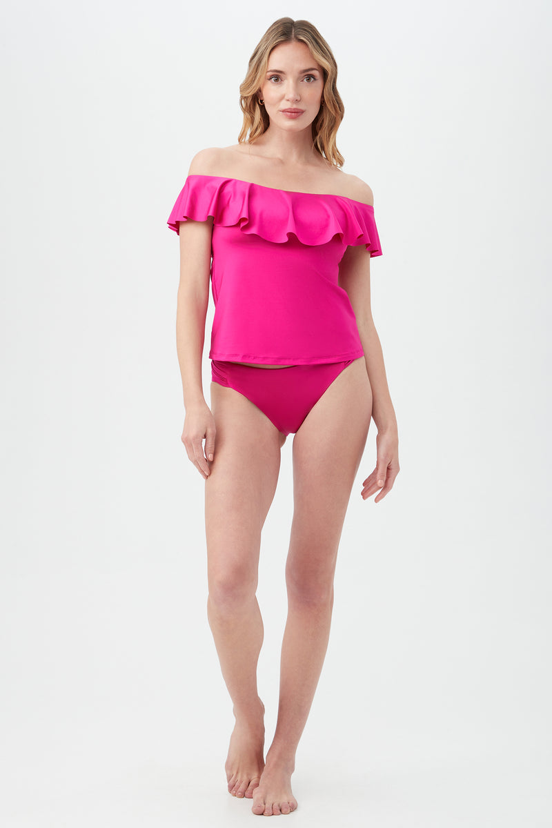 A woman stands barefoot against a white background wearing the Women's Monaco Off The Shoulder Ruffle Tankini Swim Top in bright pink with matching bikini bottoms. She has long, wavy hair and a neutral expression.