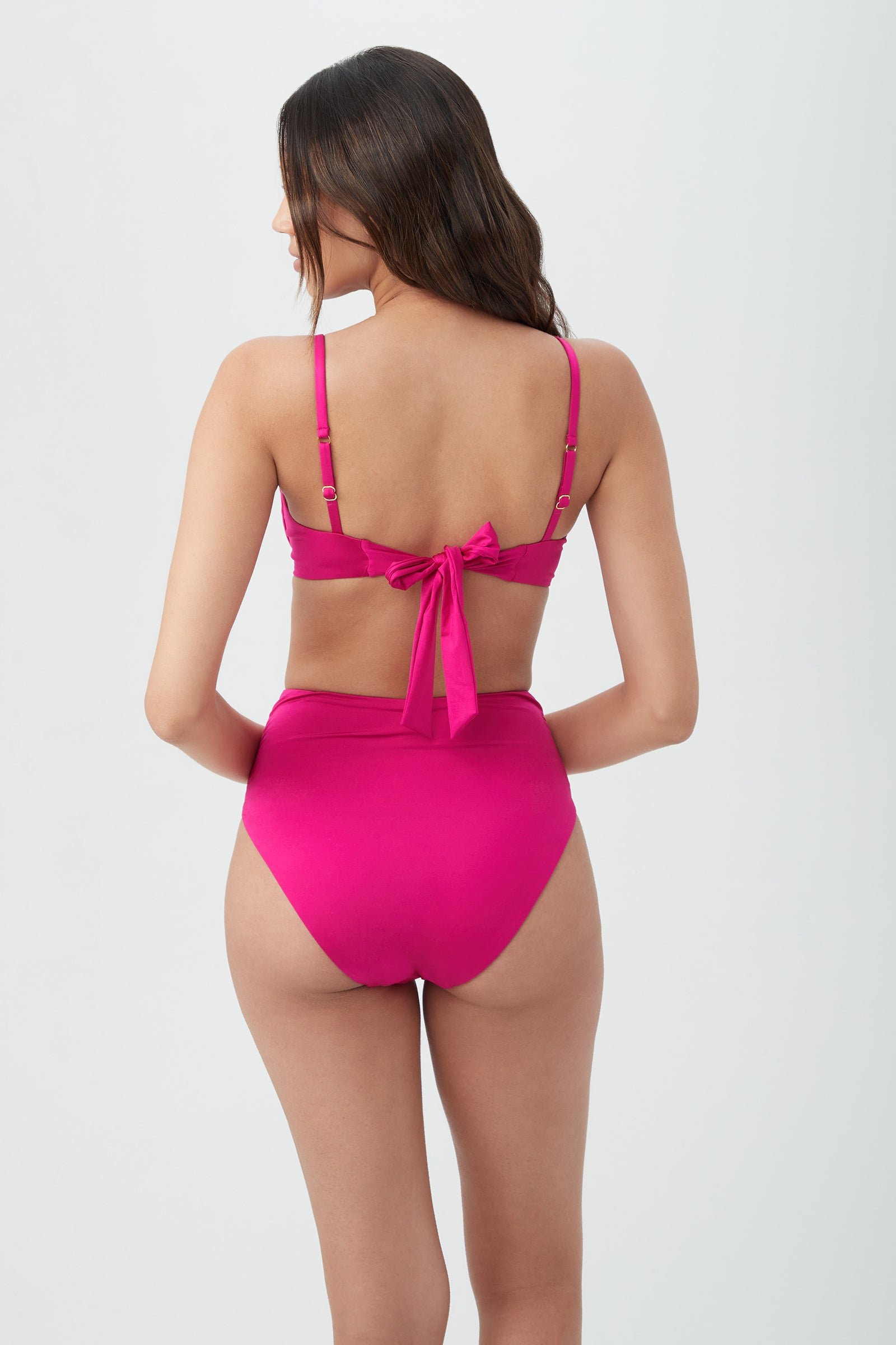 Woman with long brown hair wears the Women's Monaco Twist Front Underwire Bra Swim Top in bright pink, featuring thin adjustable straps and a back bow. Shown from behind against a plain, light background.
