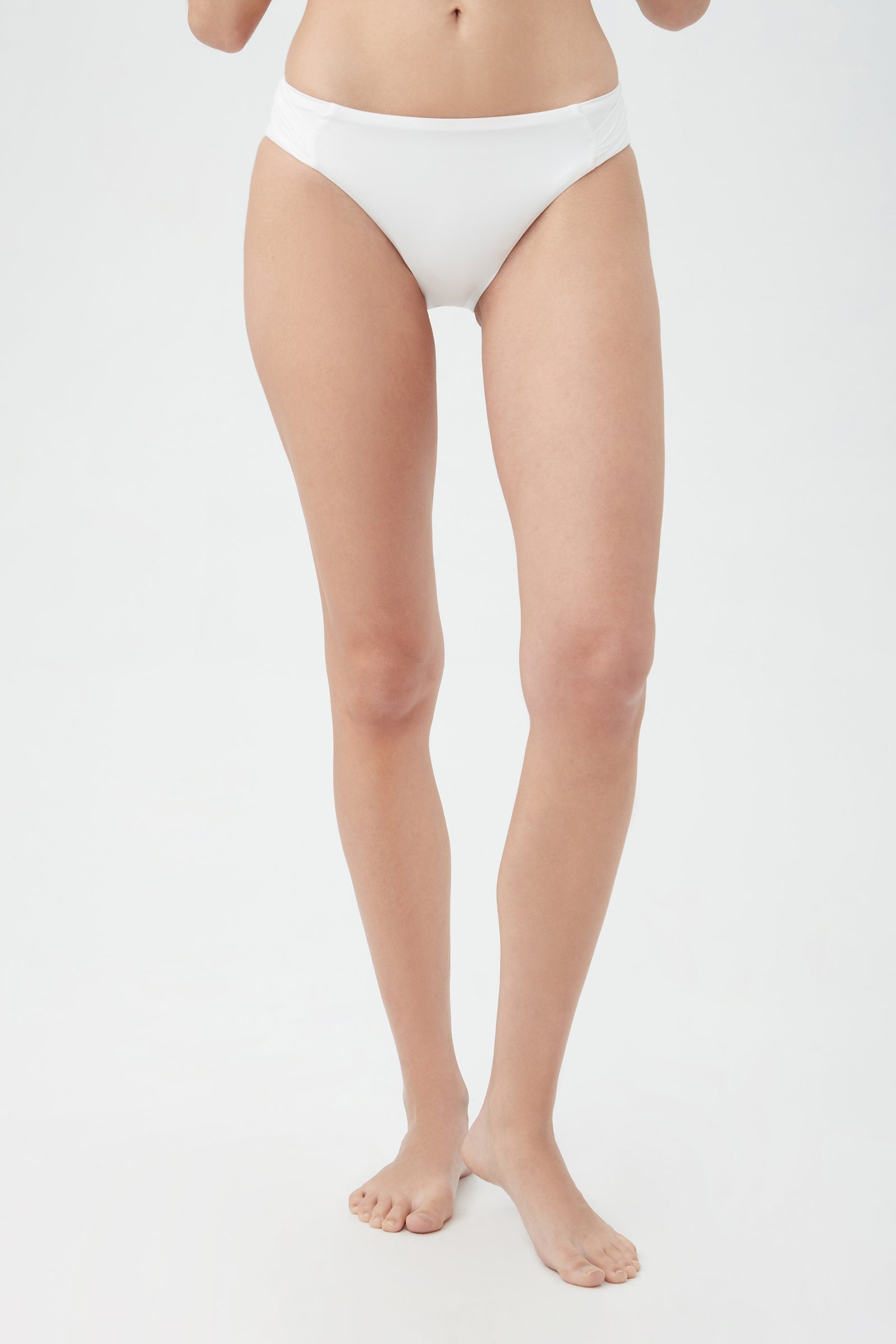 A woman stands barefoot on a white background, modeling the Women's Monaco Shirred Hipster Swim Bottom in white. Only her lower body is shown; her upper body and head are not visible.