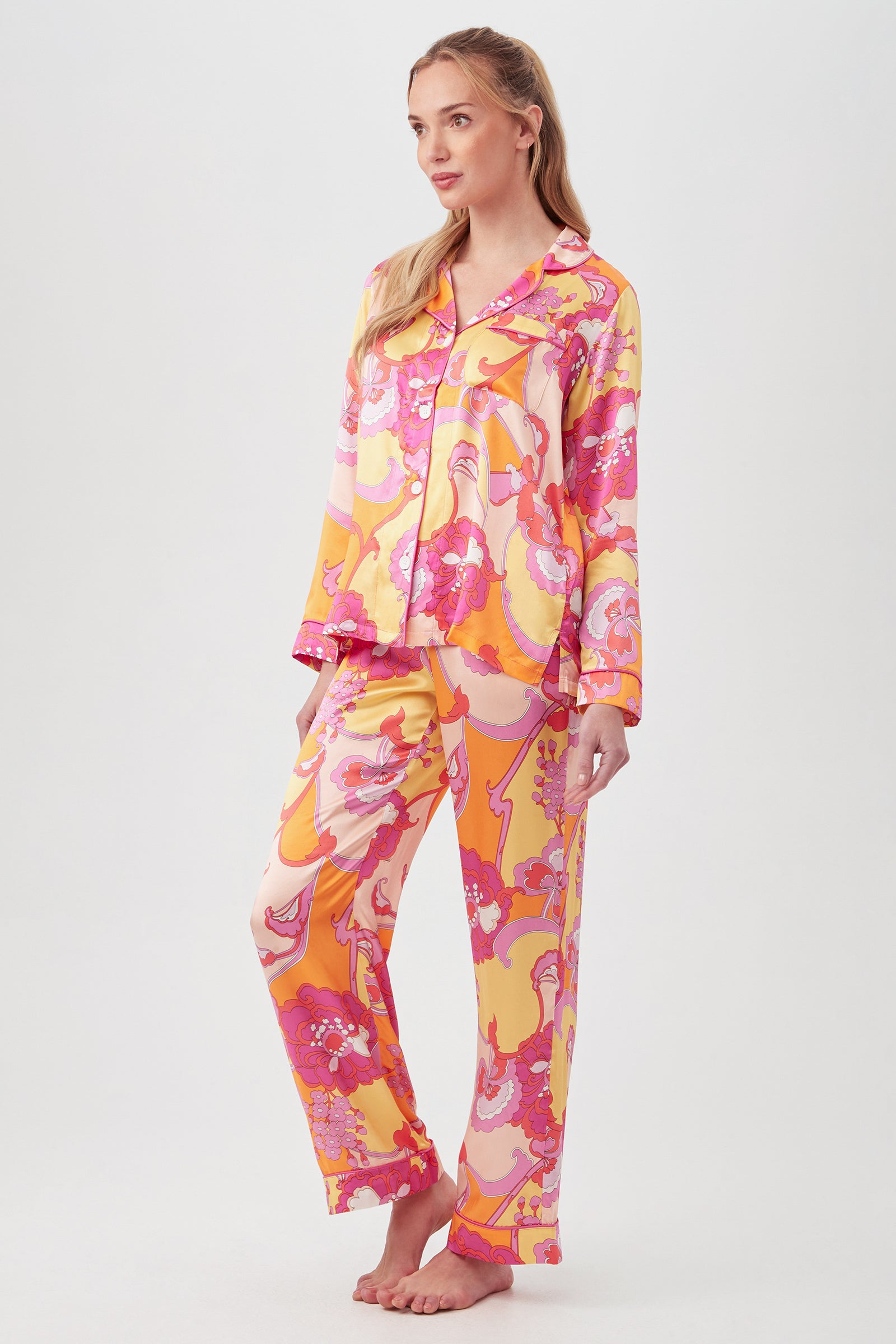 A woman wears the Apache Bloom Womens Long Sleeve Long Pant Silk PJ Set featuring a swirling pink, yellow, and orange pattern. She stands facing forward with a slight smile against a plain white background.