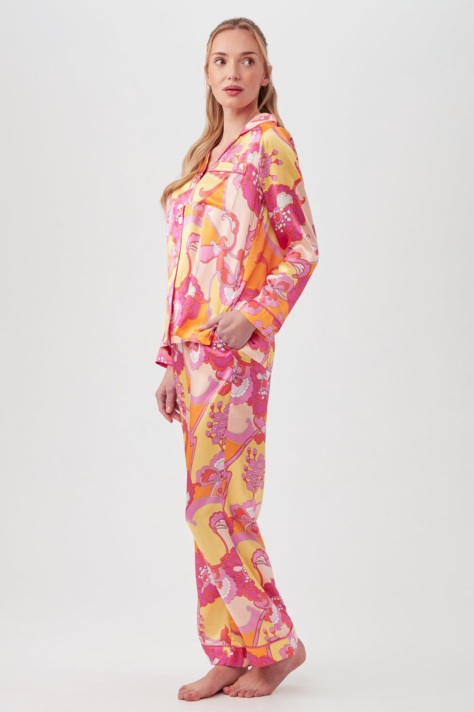 A woman stands barefoot in the Apache Bloom Womens Long Sleeve Long Pant Silk PJ Set, featuring a vibrant pink, yellow, and orange abstract floral print, posing against a plain white background.