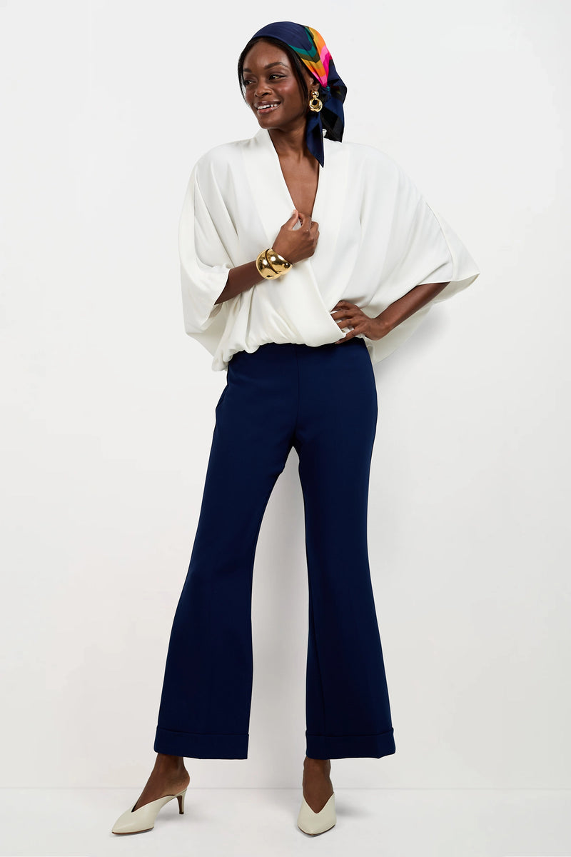A woman stands against a white background wearing the Anora V-Neck Wrap Top with wide sleeves, navy wide-leg pants, white heels, gold bracelets, large earrings, and a colorful scarf tied around her head.