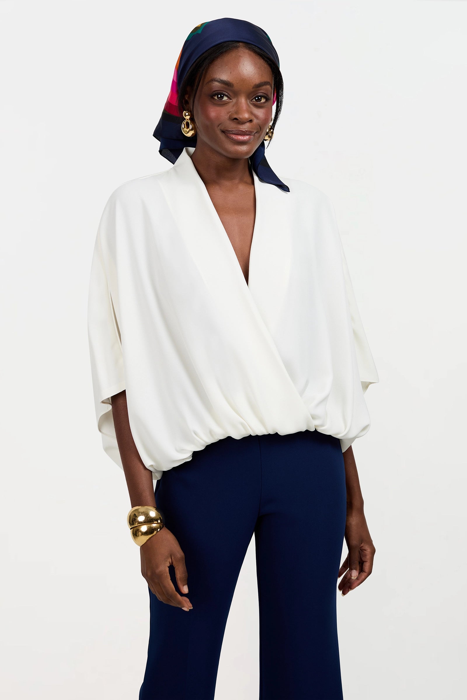 A woman wears the Anora V-Neck Wrap Top, styled with navy blue pants, a vibrant headscarf, hoop earrings, and gold bangles. She stands before a plain white background with a gentle smile.