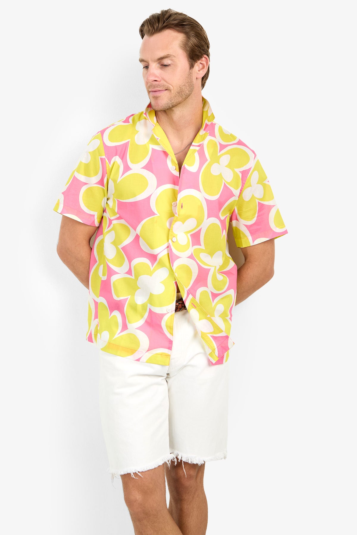 A man wearing the Felix Camp Shirt, featuring pink, yellow, and white florals, and white shorts stands against a plain background with arms relaxed and one leg crossed.