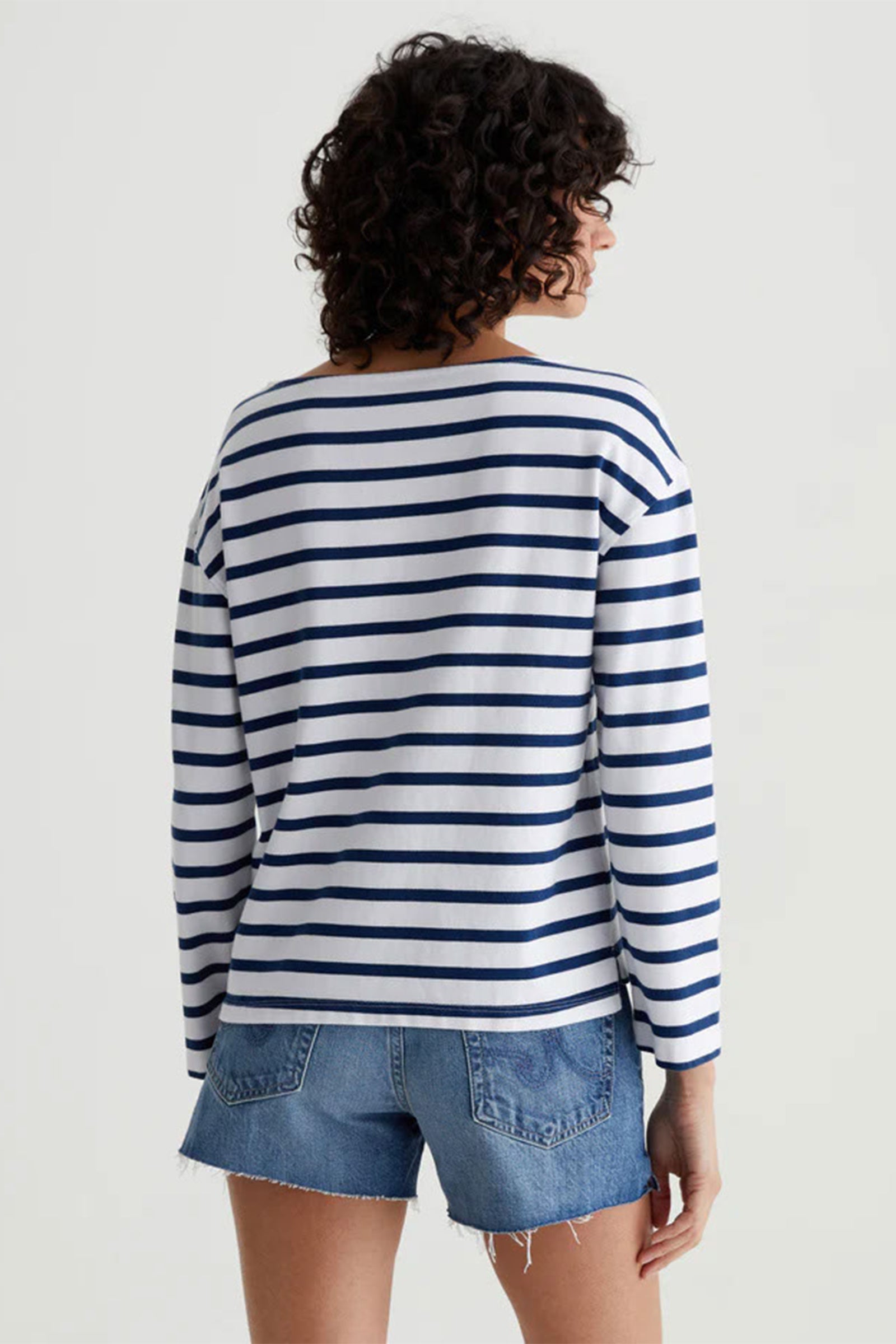 Person with curly hair, wearing the AG Womens Annie Striped Long Sleeve Top and blue denim shorts, stands with their back to the camera against a plain light background.