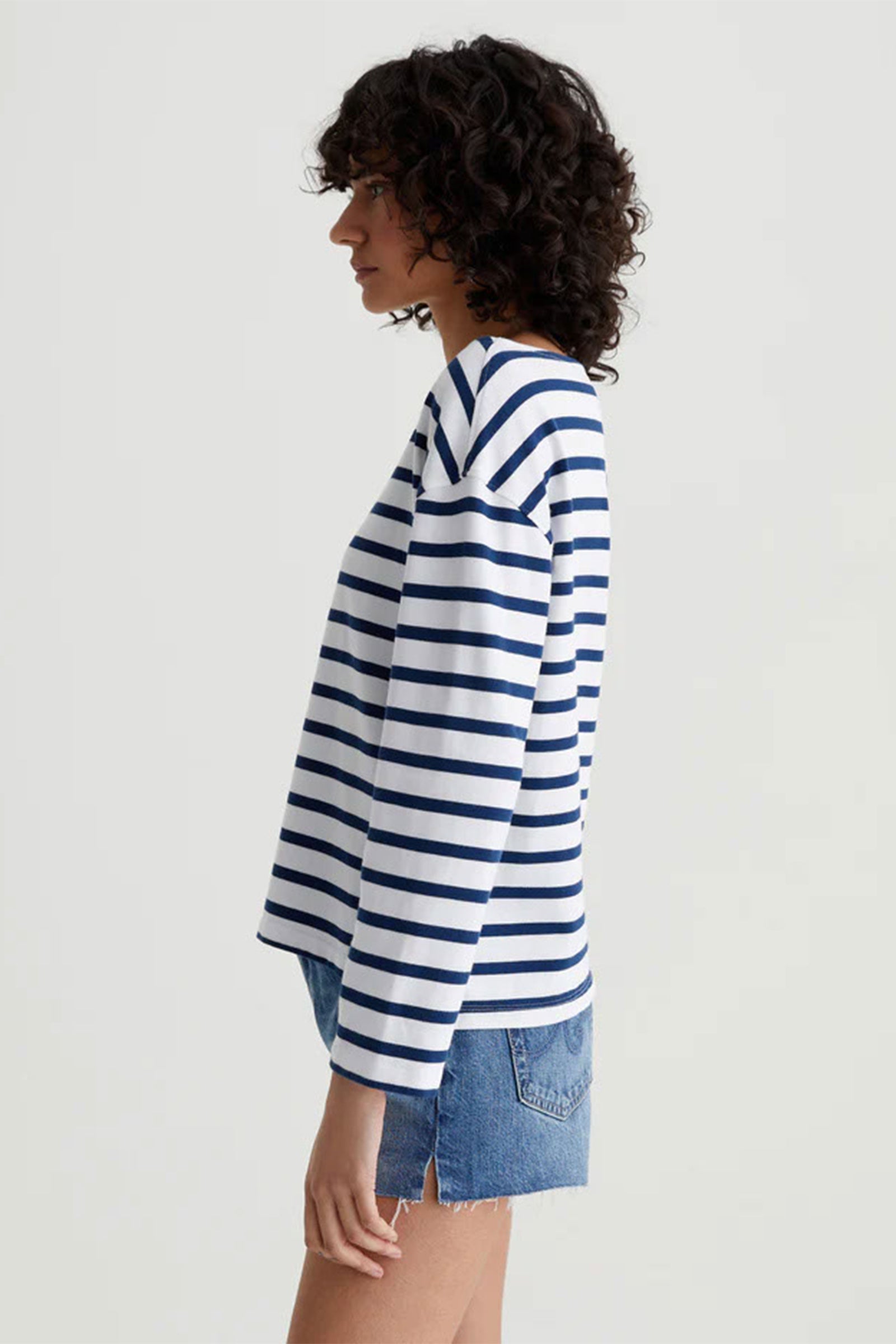 A person with curly dark hair stands in profile against a plain light background, wearing the AG Womens Annie Striped Long Sleeve Top with navy blue stripes and blue denim shorts.