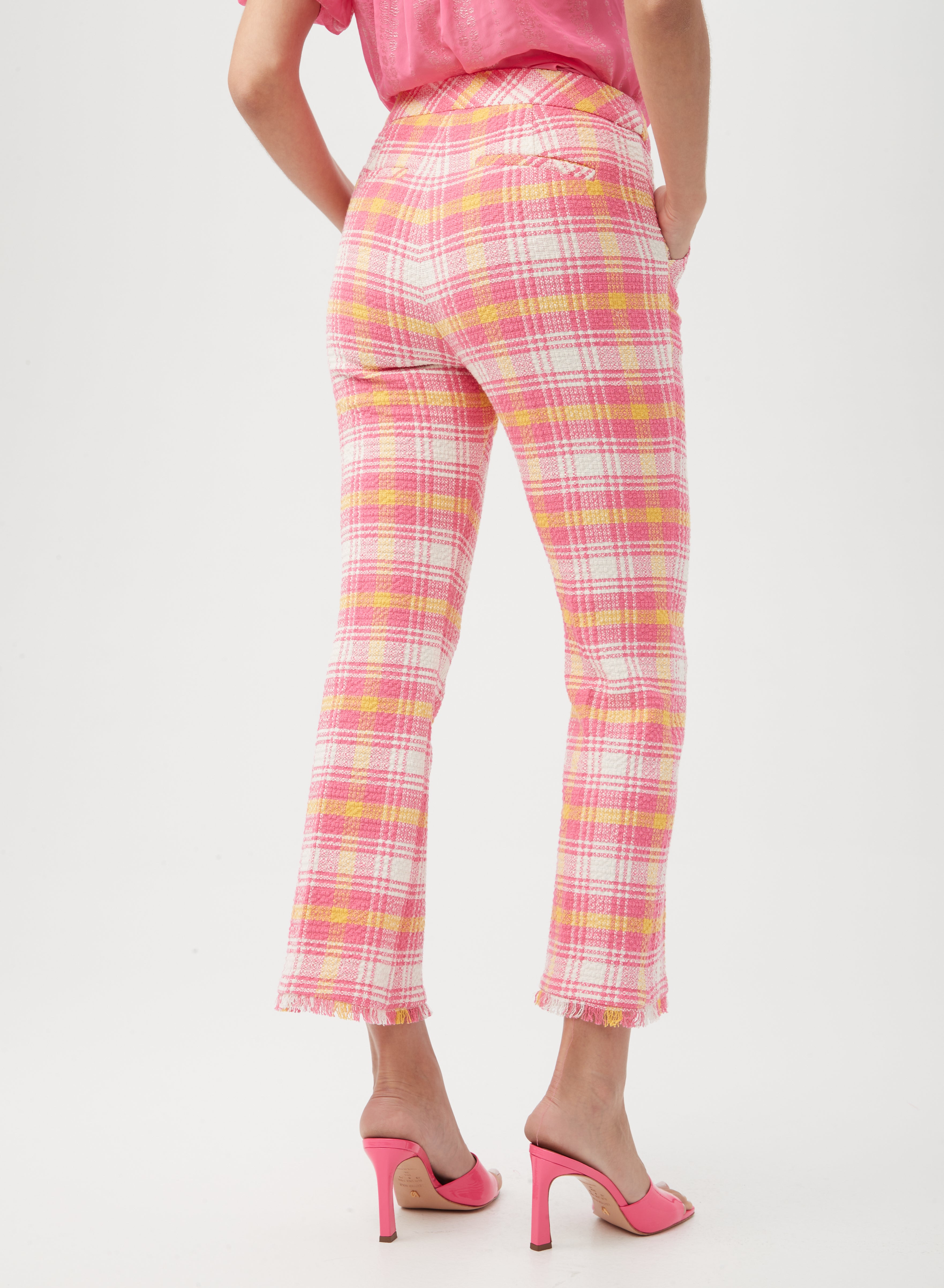A person stands facing away against a plain white background, wearing the Lulu Fringe Pant in pink and yellow plaid with frayed hems, paired with a pink shirt and pink high-heeled shoes.