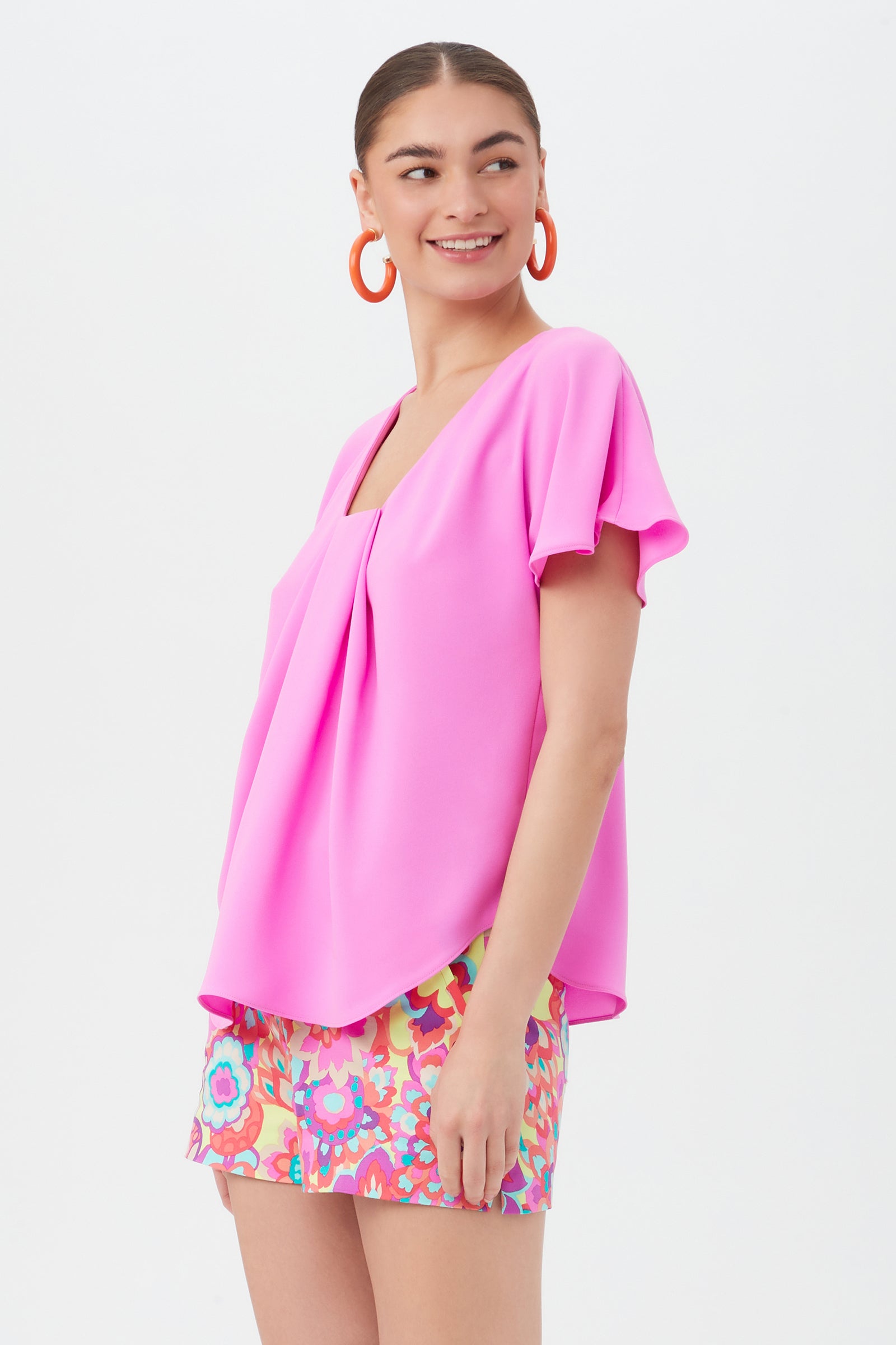A woman smiles in the Limoncello Top, a bright pink, loose-fit blouse with short sleeves, paired with multicolored patterned shorts. She wears large orange hoop earrings and has her hair pulled back against a plain white background.