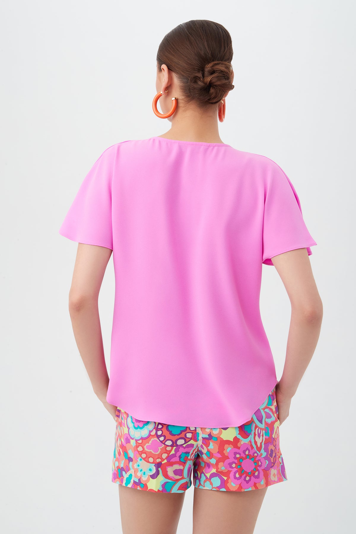 A woman with brown hair in a low bun is seen from the back, wearing the bright pink Limoncello Top, colorful patterned shorts, and large orange hoop earrings against a plain white background.