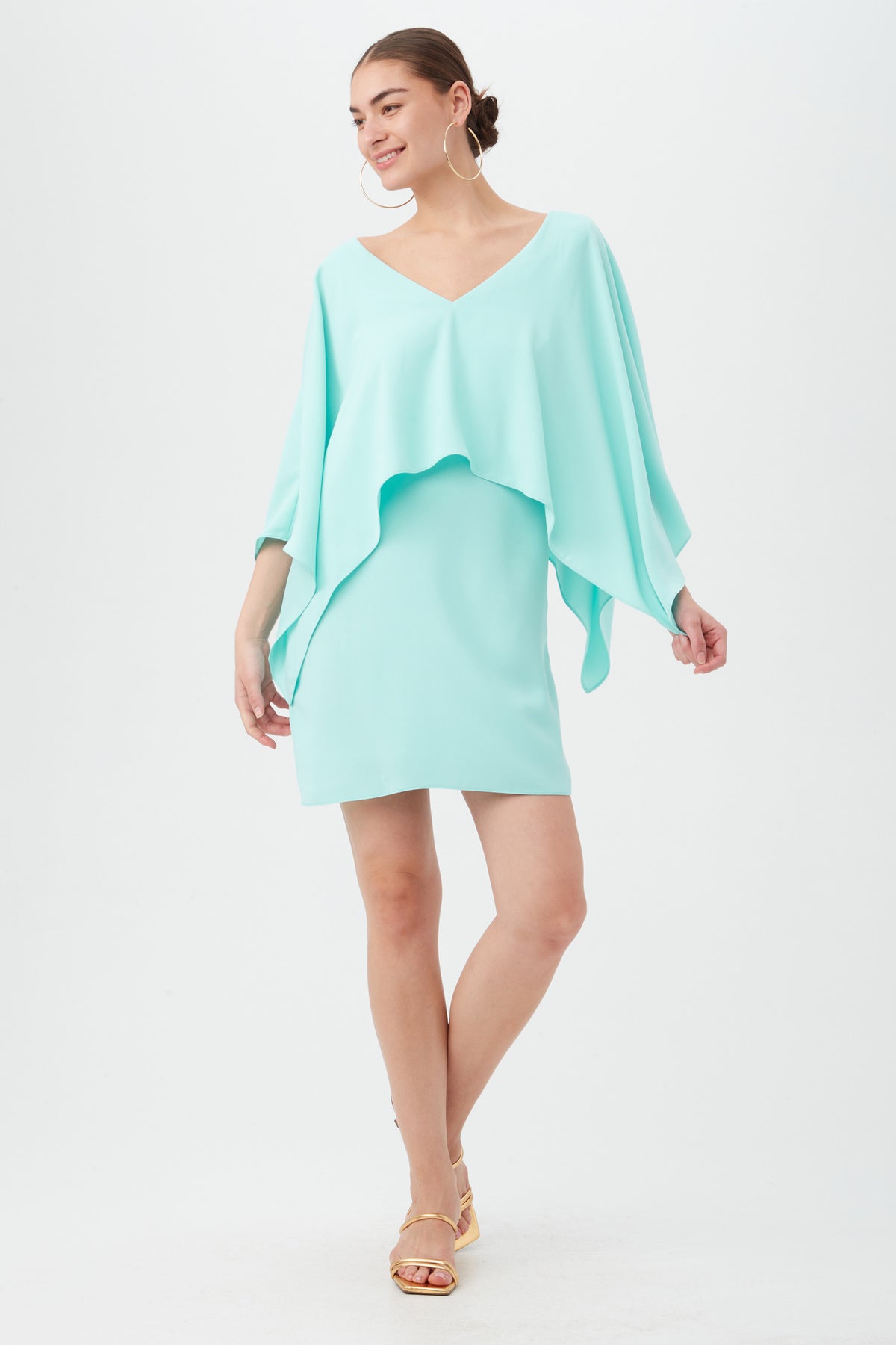 A woman wearing the Azzurra Dress in Marina, featuring a flowy, asymmetrical silhouette with wide sleeves and a V-neckline, smiles against a plain white background. She pairs it with gold sandals and hoop earrings.