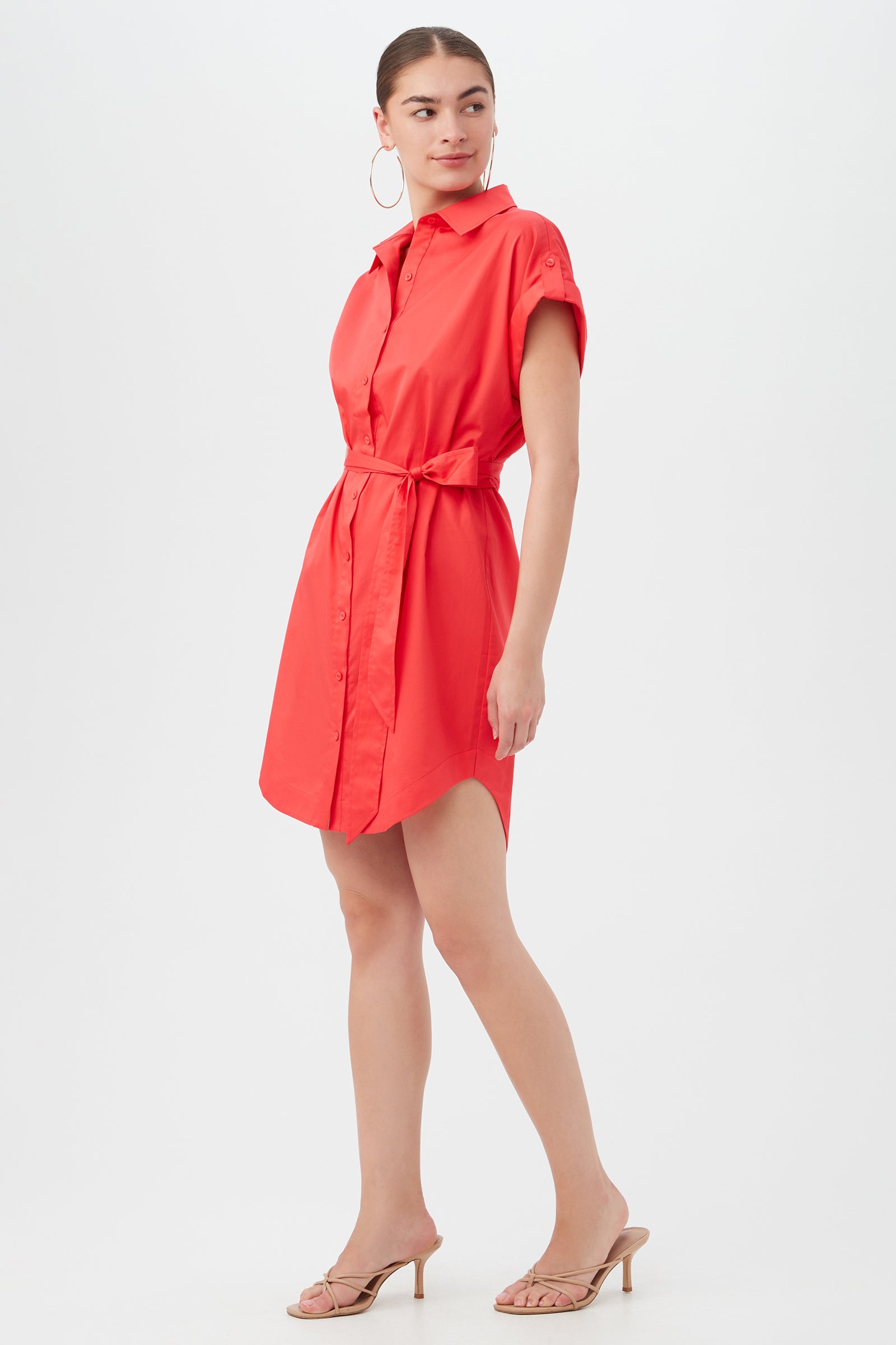 A woman stands sideways and looks over her shoulder, wearing the Simpatico Dress—a bright red, short-sleeve button-down shirt dress with a tie belt—paired with hoop earrings and heeled sandals against a plain white background.