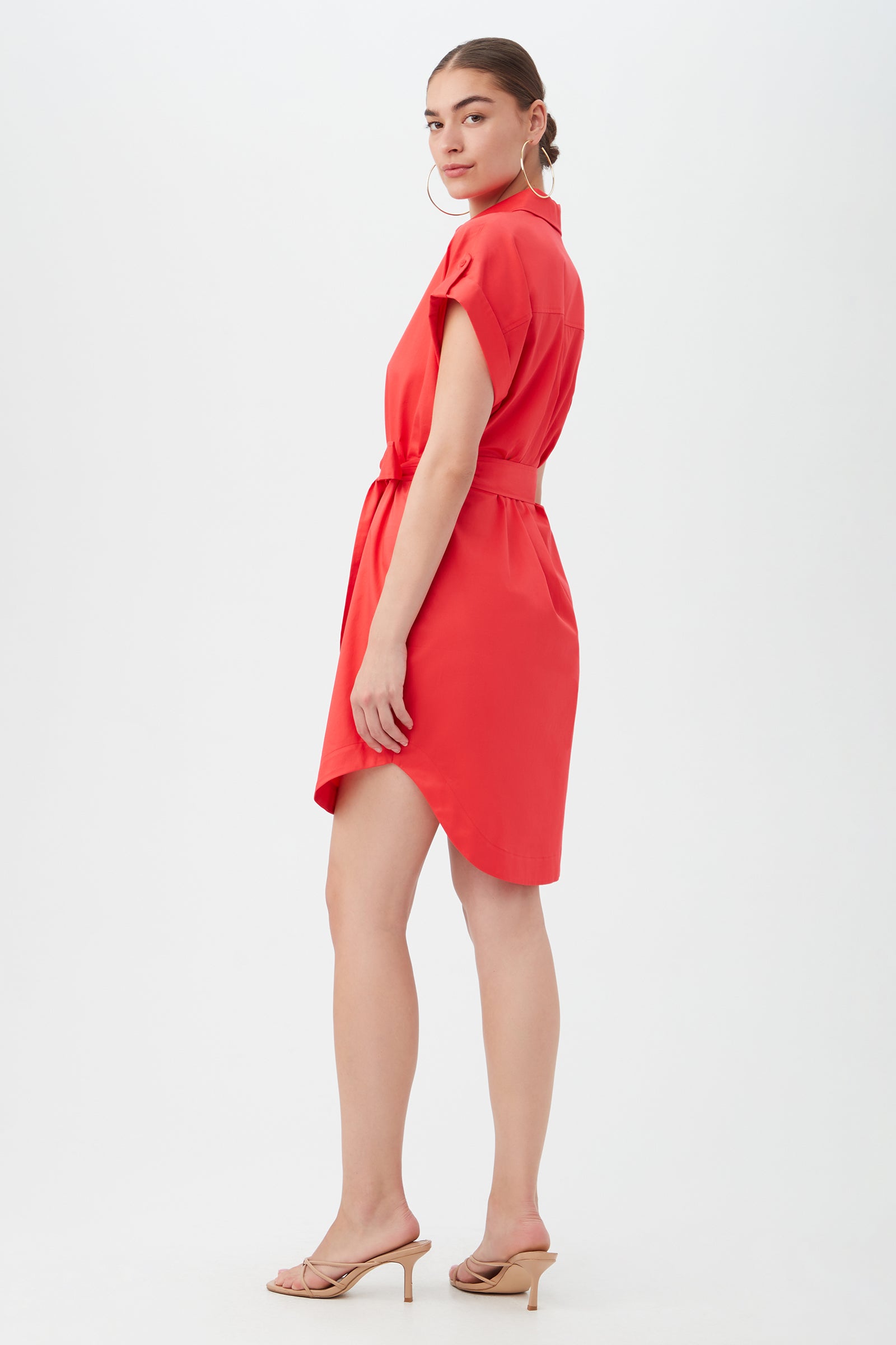 A woman models the Simpatico Dress—red, short-sleeve, and belted—paired with beige heeled sandals. She stands against a plain white background, facing slightly away while glancing over her shoulder.