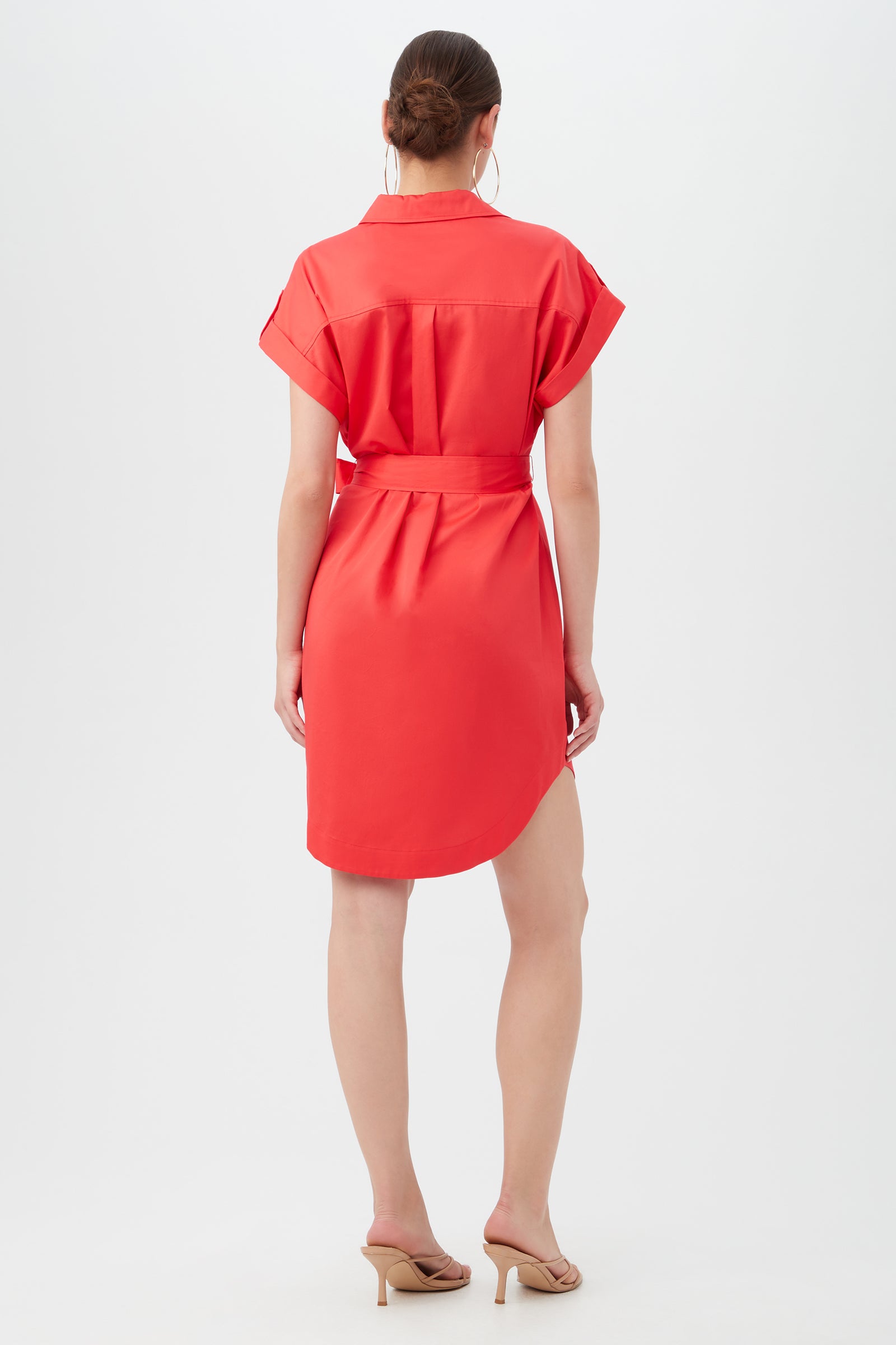 A woman stands facing away, wearing the short-sleeved, belted red Simpatico Dress and beige sandals. Her hair is tied back in a neat bun as she poses against a plain white background.