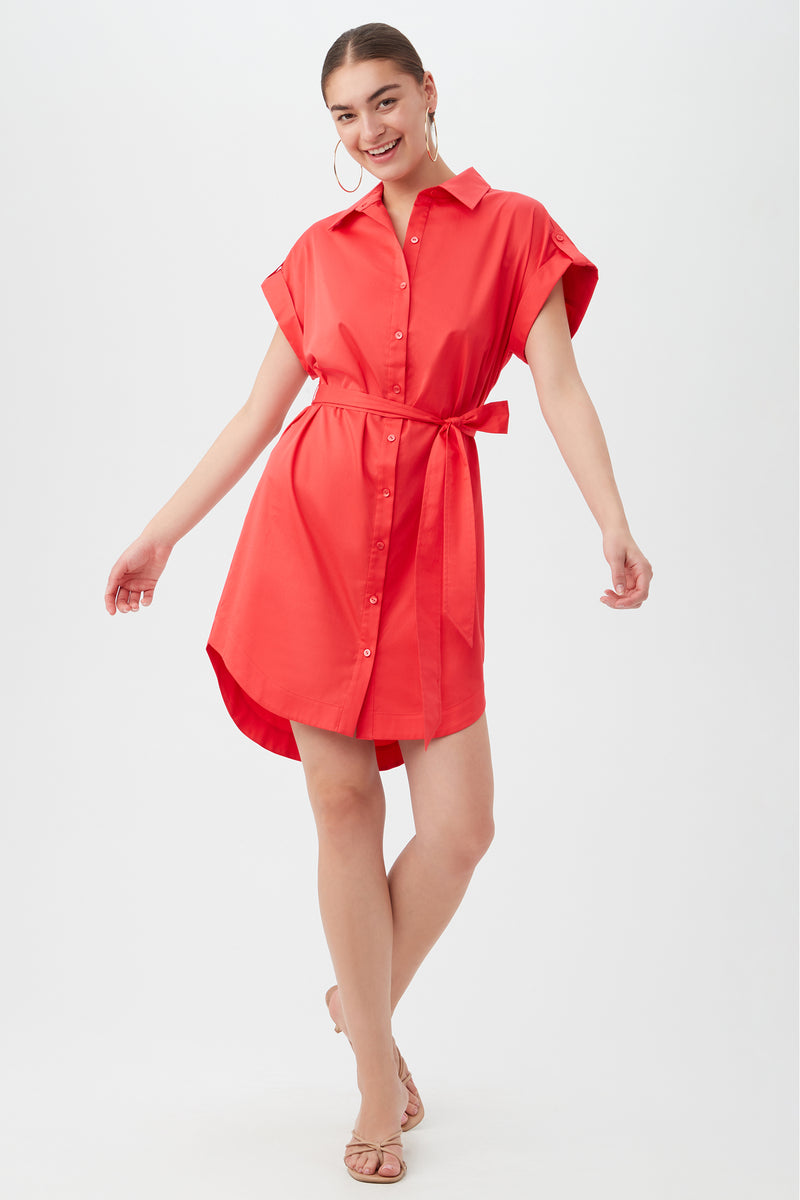 A woman wears the Simpatico Dress in Capri Coral, featuring short sleeves, a collar, button front, and waist tie, smiling and posing with one foot slightly lifted against a plain light background.