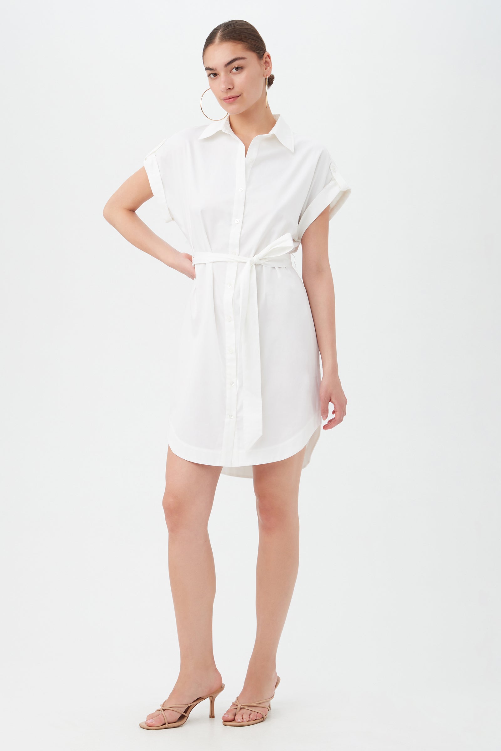 A woman poses with one hand on her hip and a slight smile, wearing the Simpatico Dress—a short-sleeve, button-down white shirtdress with a waist belt—paired with hoop earrings and nude heeled sandals against a white background.