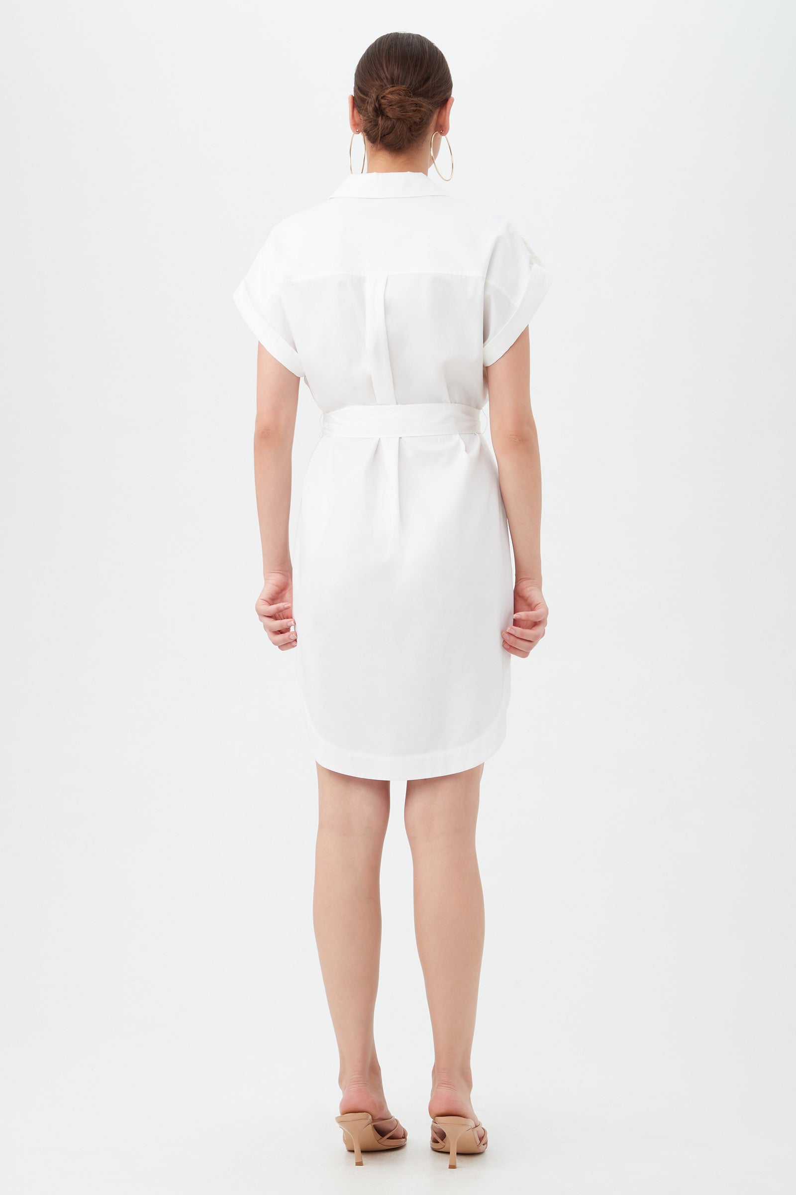 A woman with her hair in a low bun faces away, wearing the Simpatico Dress—a white, short-sleeve, knee-length belted dress—paired with beige heeled sandals against a plain white background.