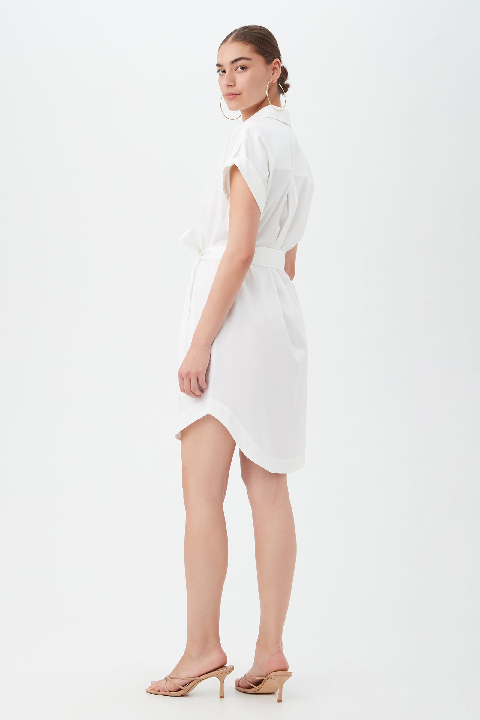 A woman with her hair tied back poses against a white background, wearing the short-sleeved, belted Simpatico Dress in white and beige heeled sandals, looking over her shoulder.