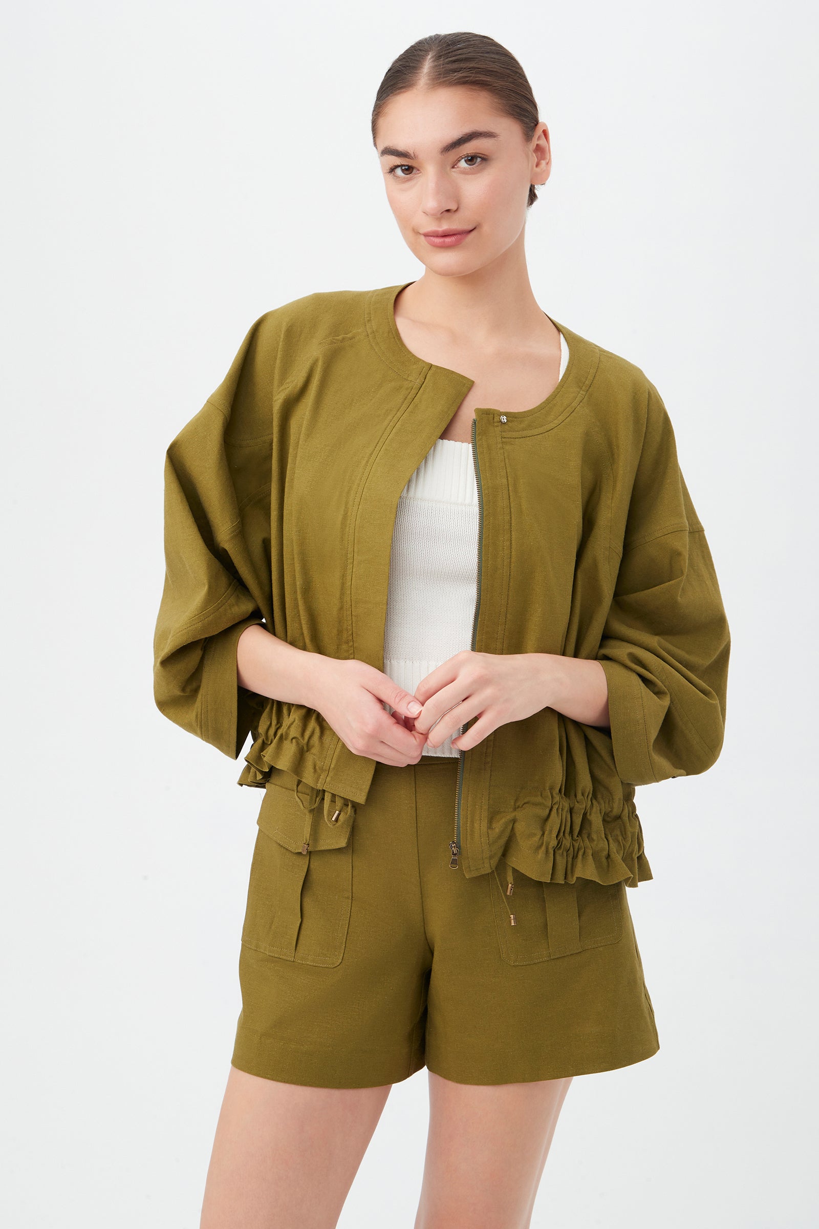 A woman stands against a white background wearing the Prita Jacket in Sorrento Sage with ruffled details, paired with matching shorts and a white ribbed top. Her hair is pulled back.