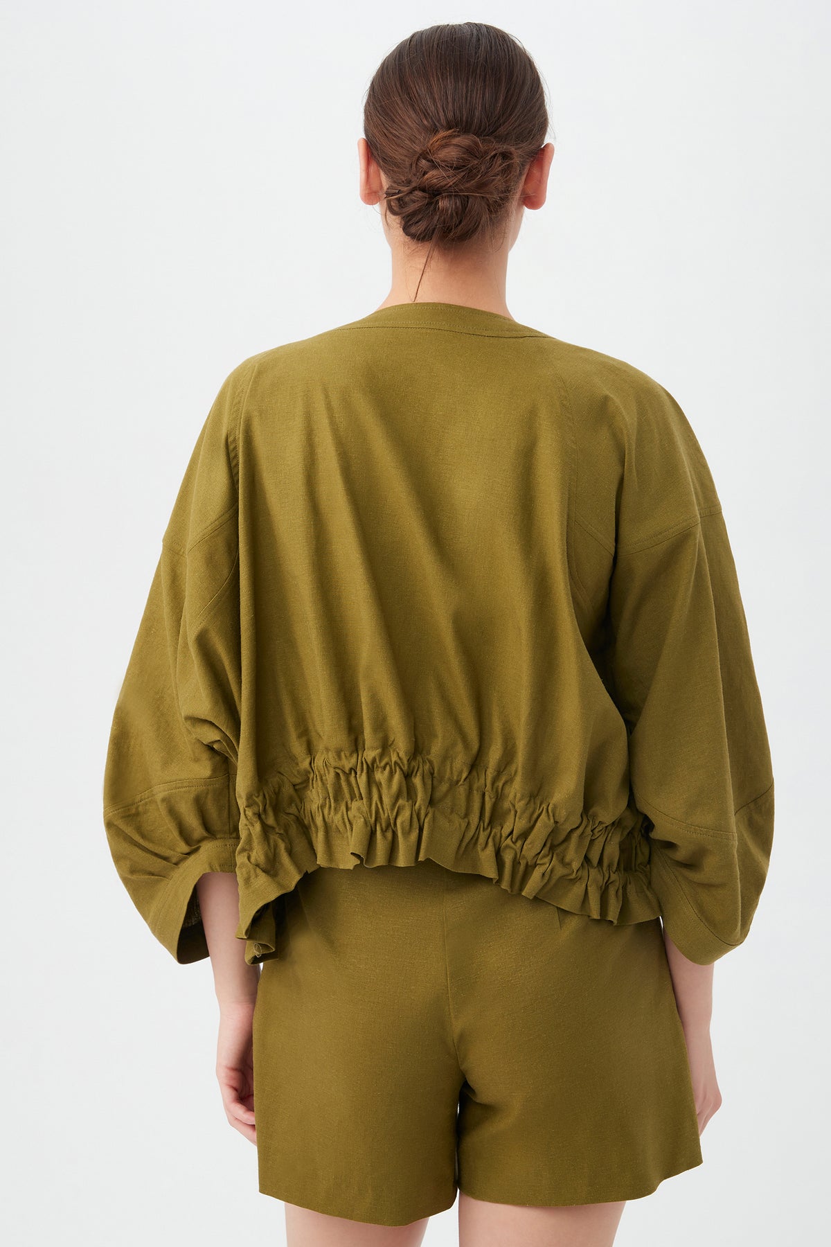 A woman with brown hair in a low bun is seen from the back, wearing the olive green Prita Jacket with a gathered waist and matching shorts, set against a plain white background.