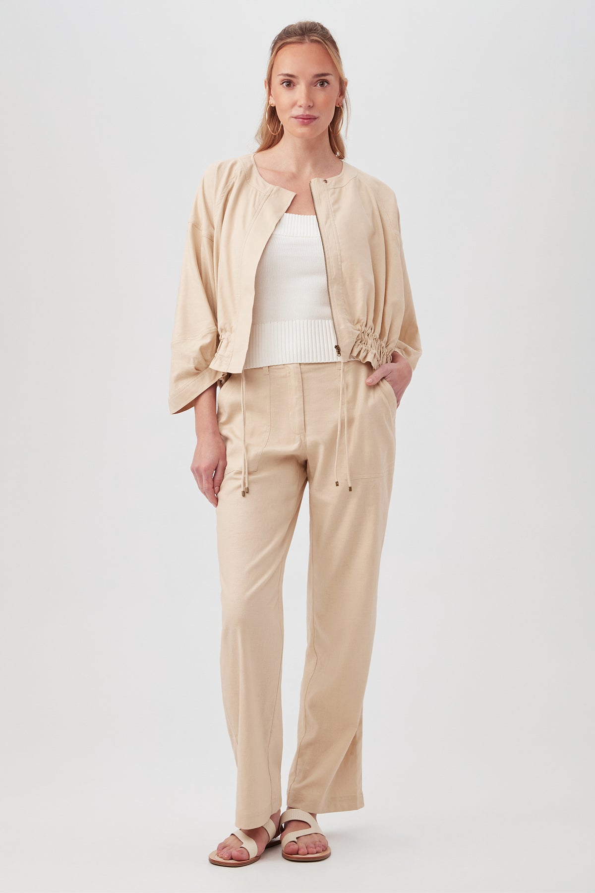 A woman stands facing forward, wearing the beige Prita Jacket with drawstrings, matching pants, a white ribbed top, and brown sandals. She has long light brown hair and a relaxed, confident expression.
