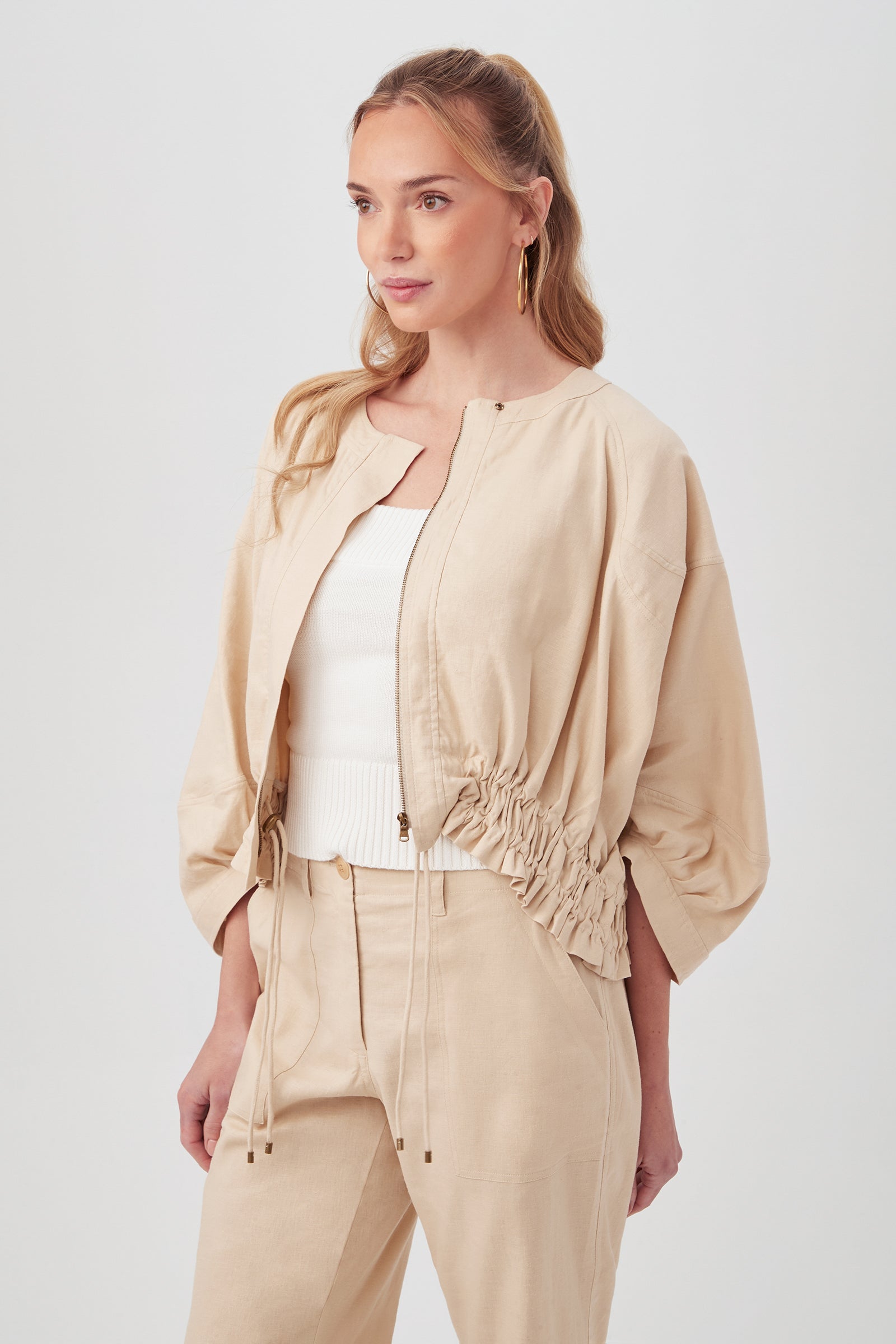 A woman with long, light brown hair models the beige Prita Jacket over a white top and matching beige pants, standing against a plain light background.