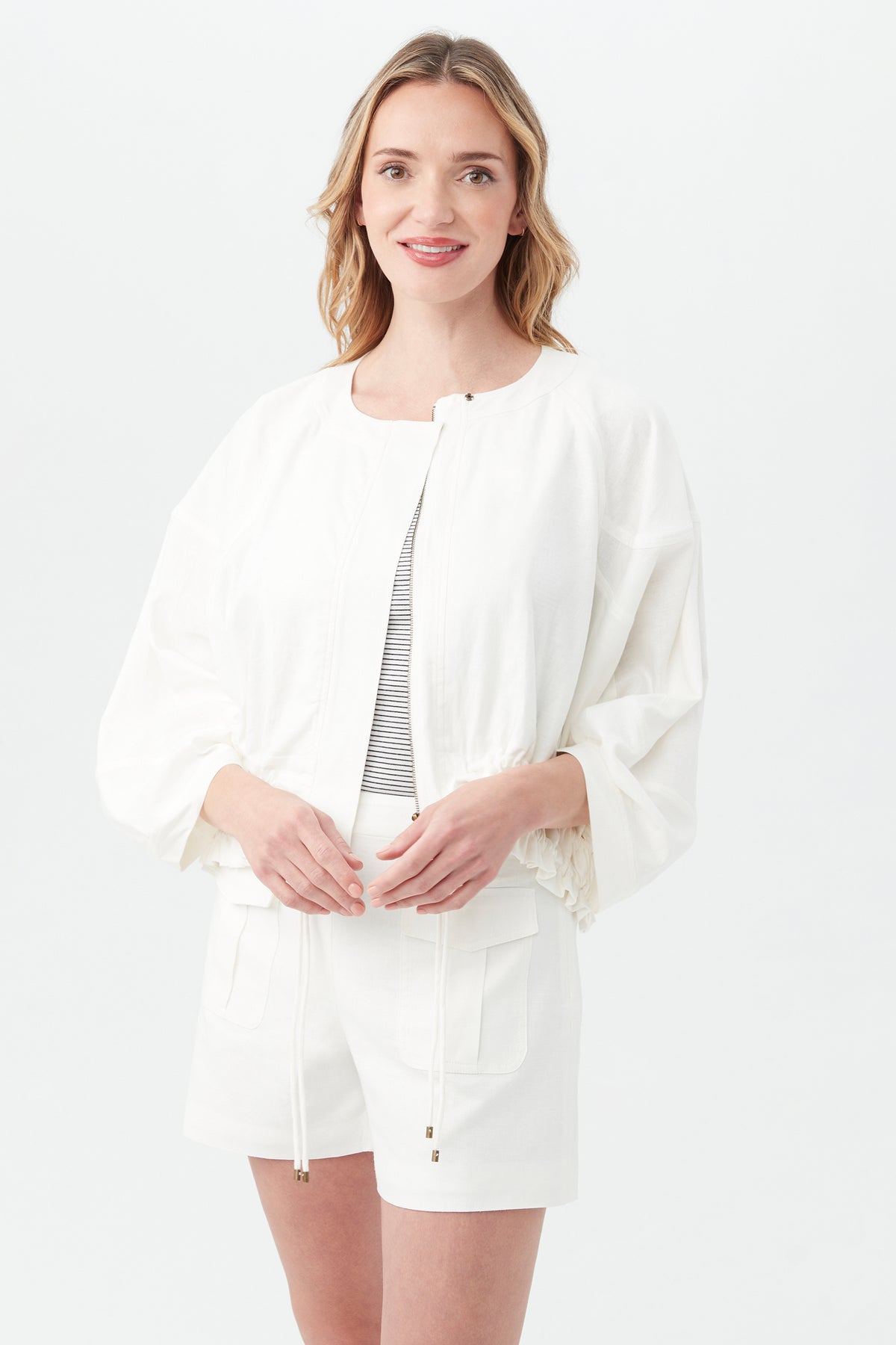 A woman with wavy blonde hair smiles as she stands against a light background, wearing the Prita Jacket in Whitewash over a striped top and white shorts.