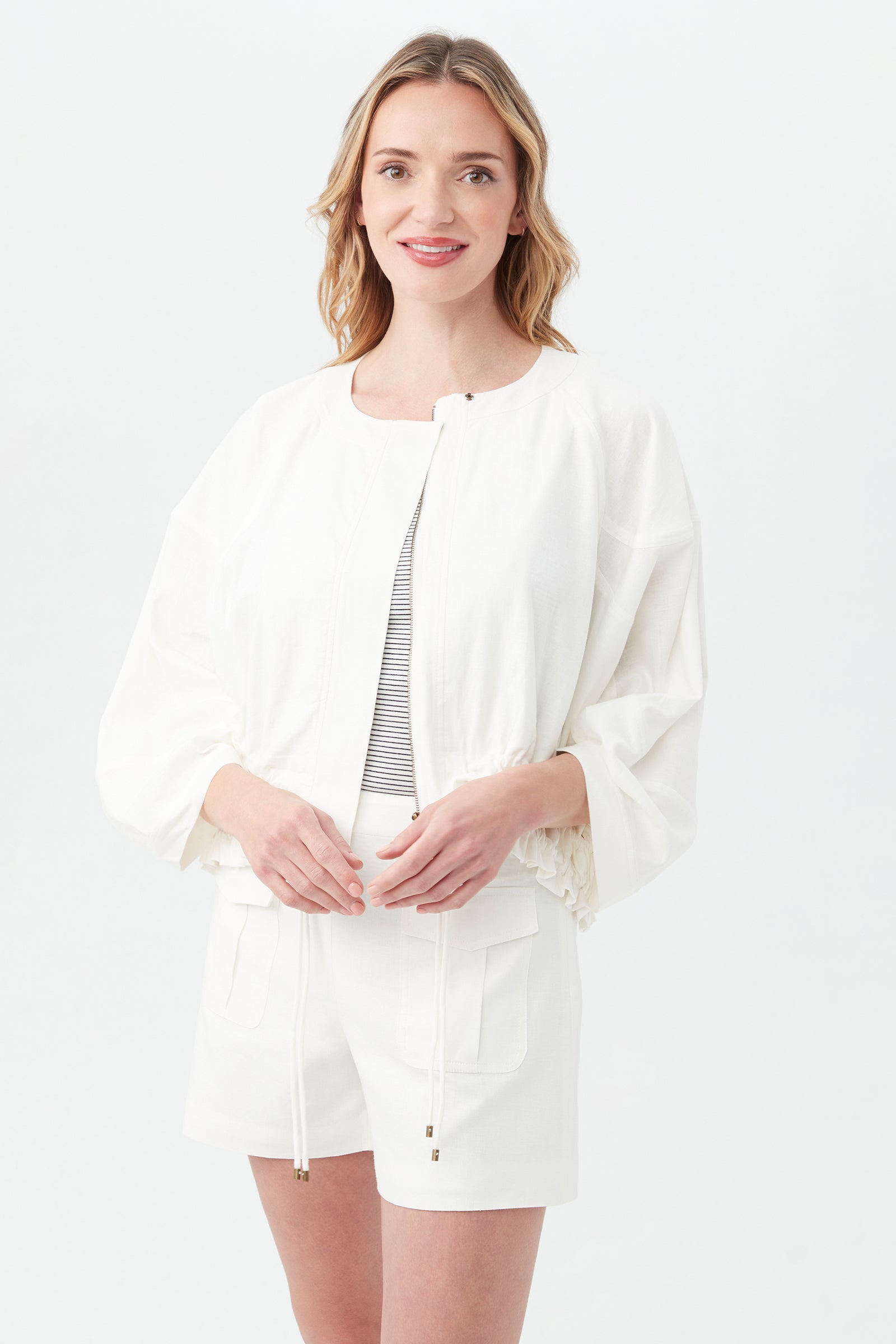 A woman with wavy blonde hair smiles as she stands against a light background, wearing the Prita Jacket in Whitewash over a striped top and white shorts.