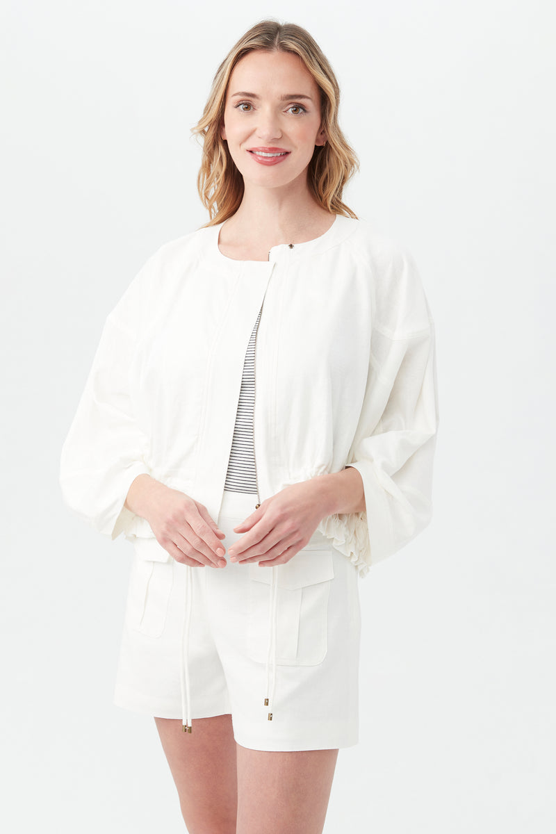 A woman with wavy blonde hair smiles as she stands against a light background, wearing the Prita Jacket in Whitewash over a striped top and white shorts.