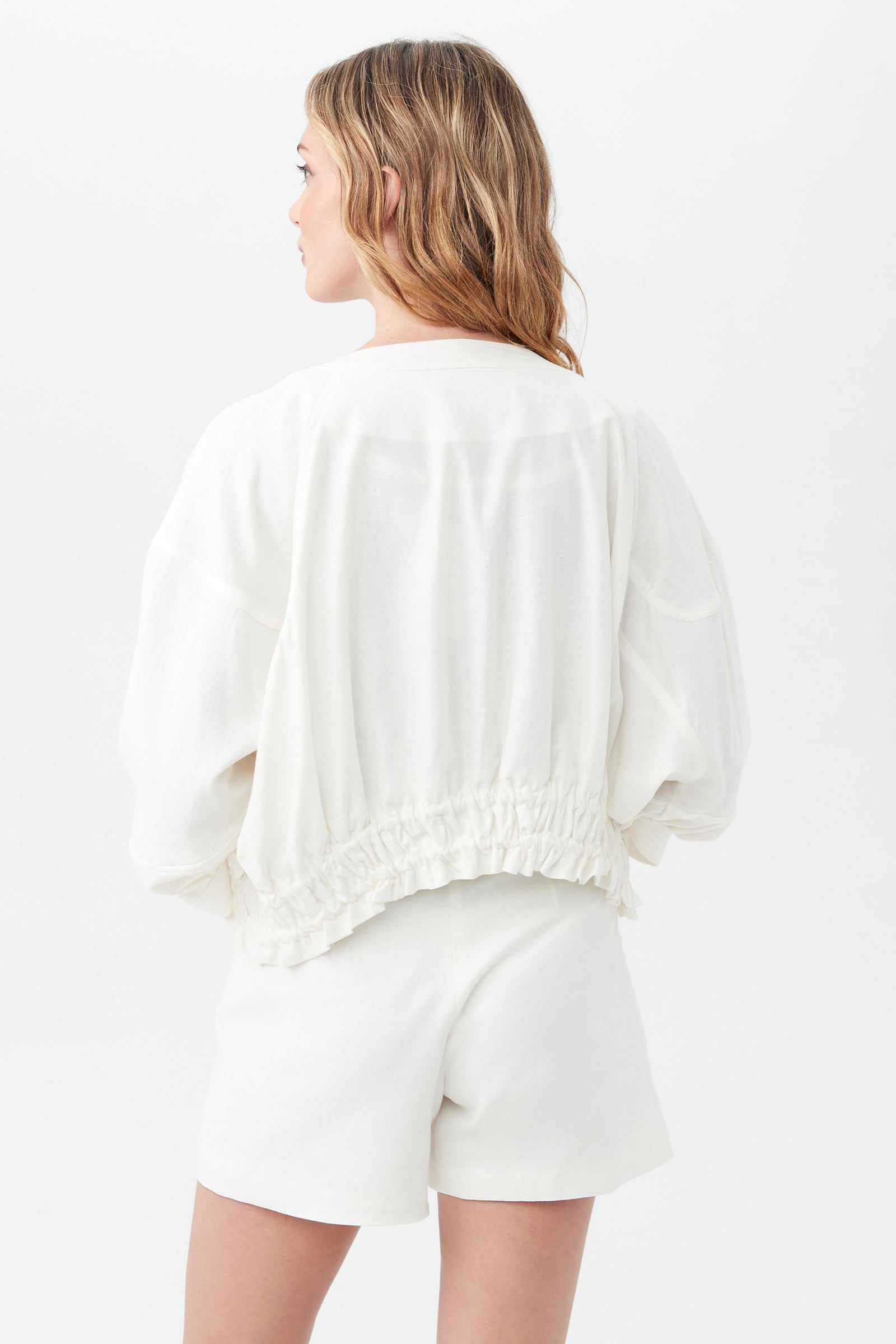 A woman with wavy, light brown hair is seen from behind, wearing the Prita Jacket—a loose white top with an elastic waistband—and matching white shorts, standing against a plain white background.