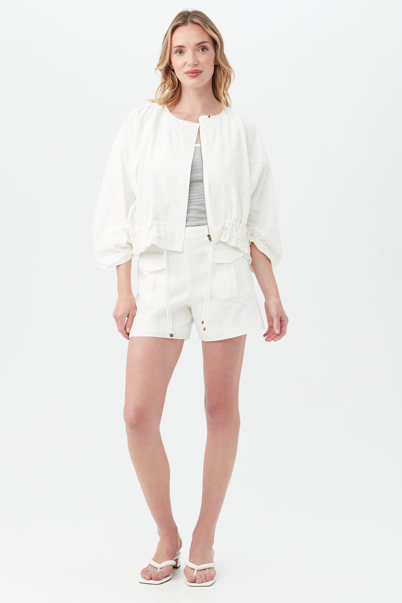 A woman with loose, wavy light brown hair and a relaxed expression wears the Prita Jacket over a light gray top, paired with white shorts and sandals, standing against a white background.