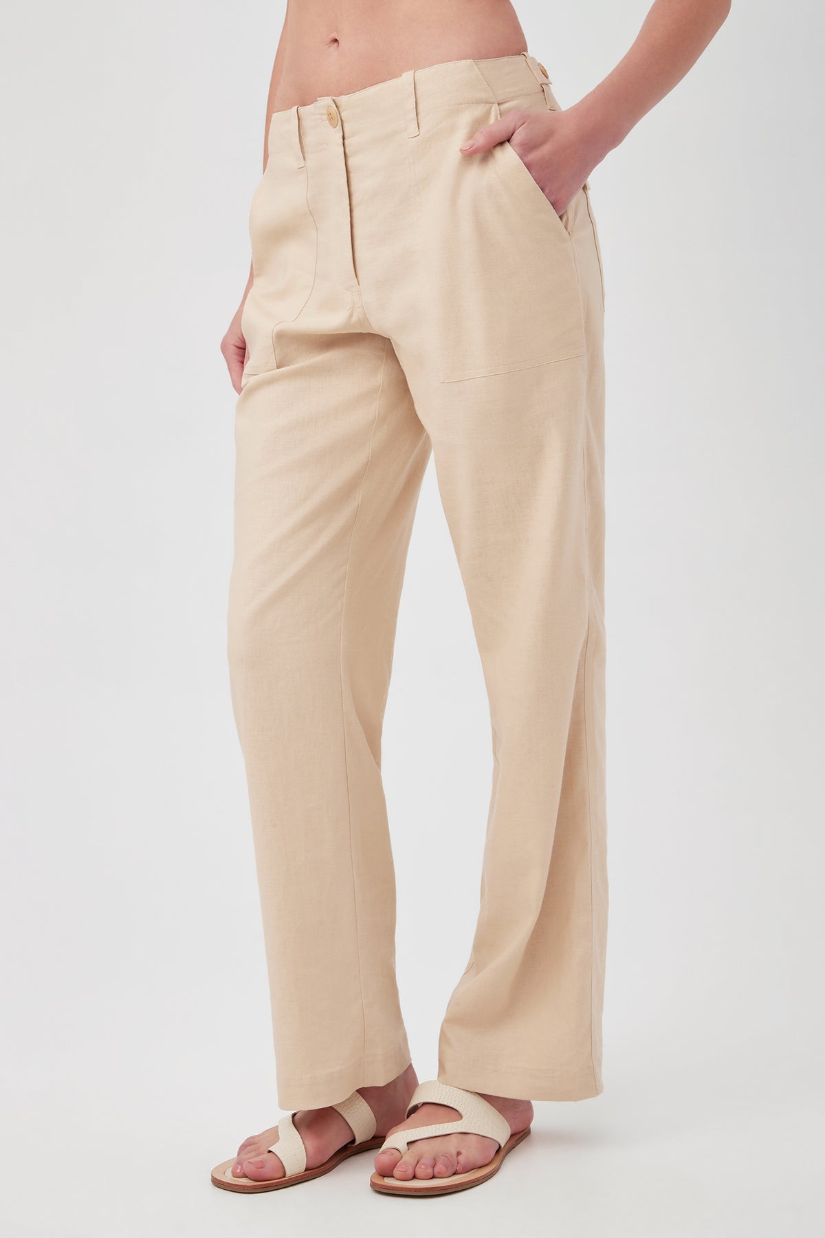 A person wears the Antonia Pant—high-waisted, beige linen pants—with hands in pockets and white sandals, standing against a plain white background. The head and upper torso are not visible.