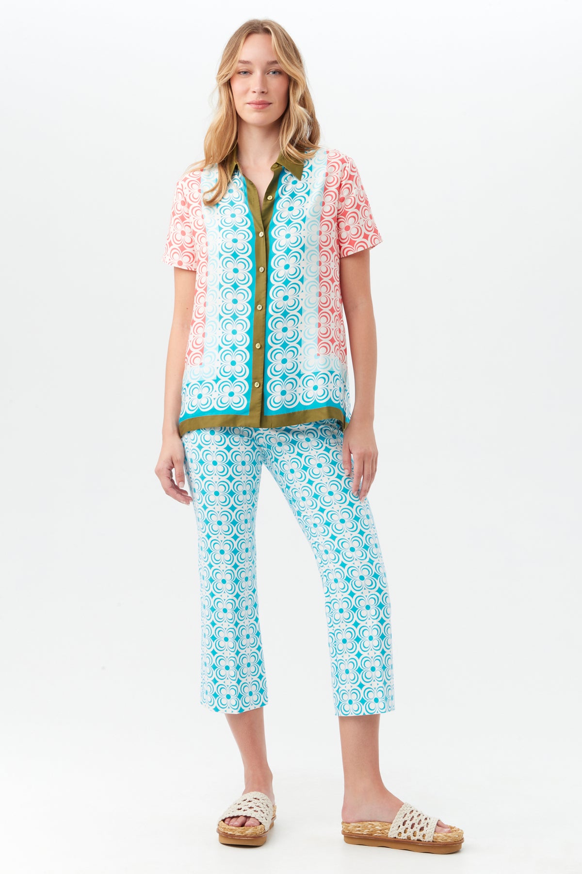 A woman with long wavy blonde hair stands against a white background wearing a short-sleeved pink and green patterned shirt, the Flaire 2 Pant in blue, and white woven sandals.