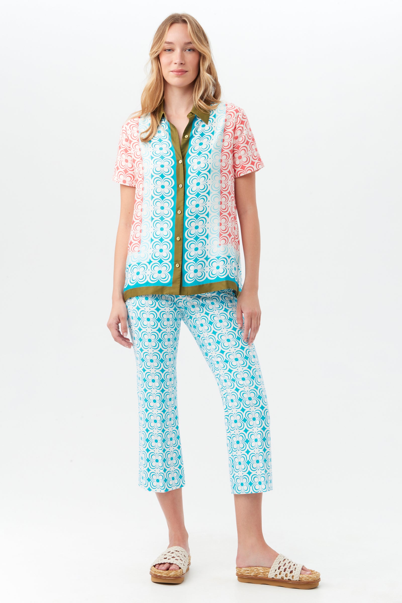 A woman with long wavy blonde hair stands against a white background wearing a short-sleeved pink and green patterned shirt, the Flaire 2 Pant in blue, and white woven sandals.