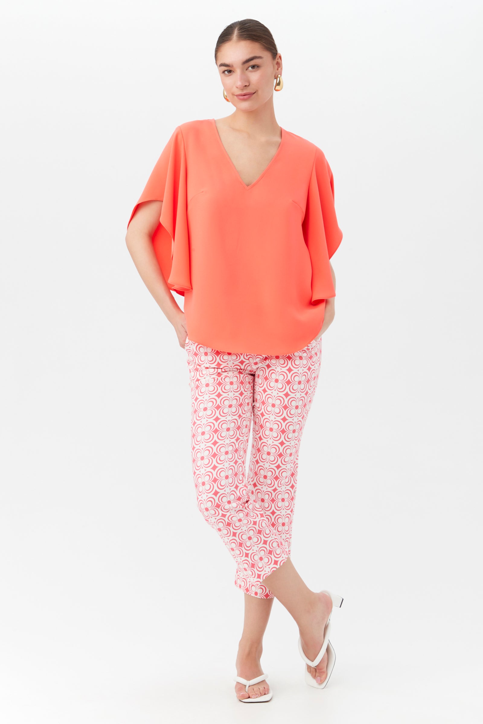 A woman stands against a white background, wearing the Flaire 2 Pant in pink and white with a coral V-neck blouse, white open-toe heels, and gold hoop earrings. She has her hands in her pockets and looks relaxed.