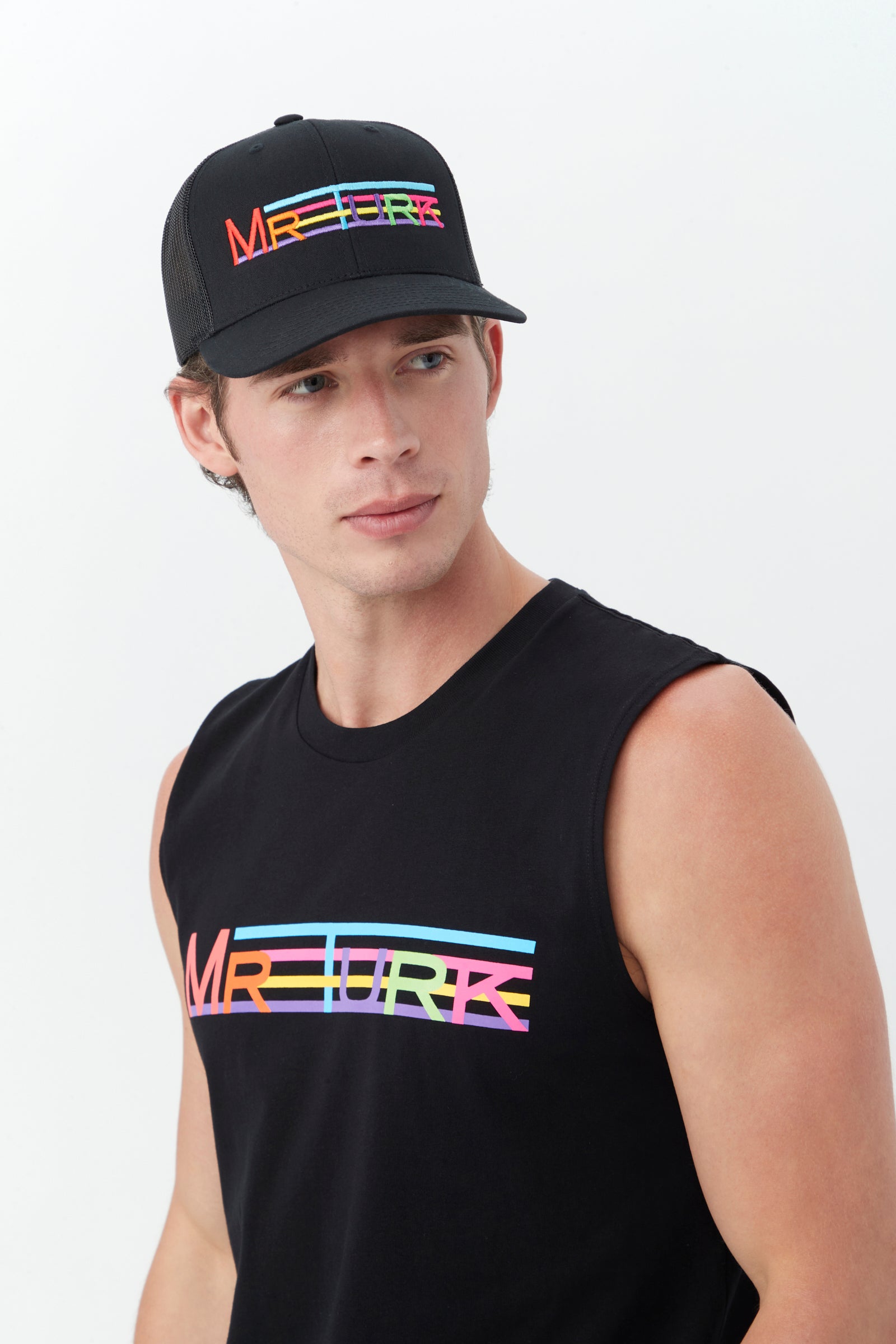 A young man wears the Mr Turk Pride Trucker Hat, a black cap with rainbow-colored MR TURK text and lines, paired with a matching sleeveless shirt, as he stands against a plain light background looking to his left.