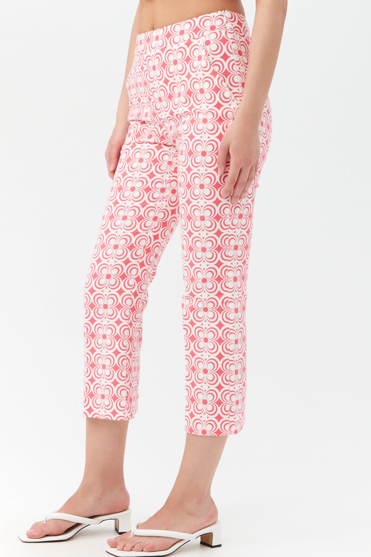 A woman wears the Flaire 2 Pant, pink and white with a geometric pattern, paired with white heeled sandals against a plain white background.