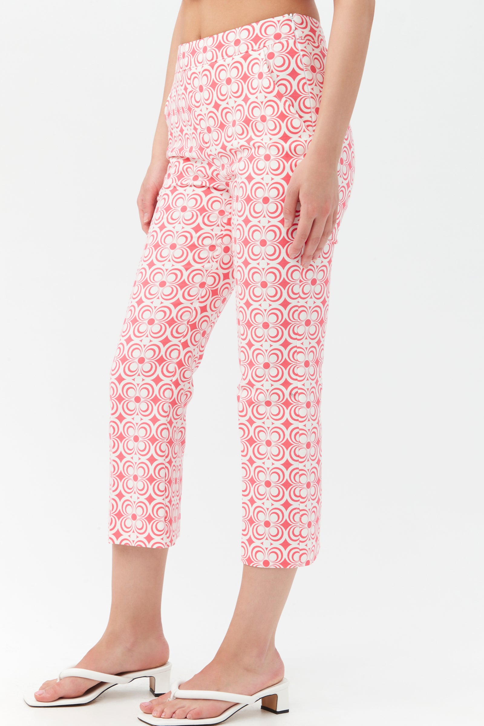 A woman wears the Flaire 2 Pant, pink and white with a geometric pattern, paired with white heeled sandals against a plain white background.