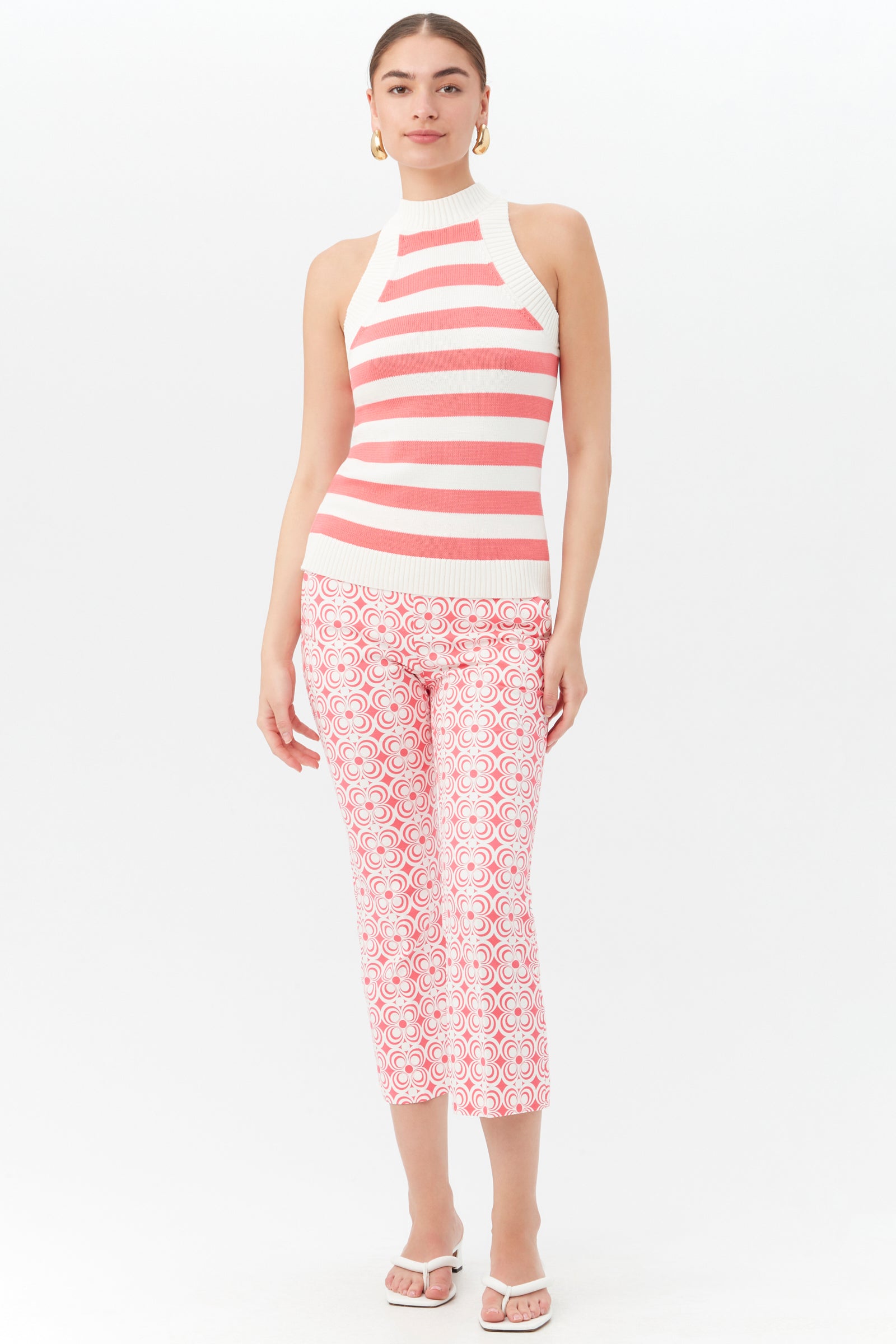 A woman poses against a white background in the Flaire 2 Pant—cropped, pink and white patterned pants—paired with a sleeveless halter-neck top, white sandals, and gold hoop earrings.