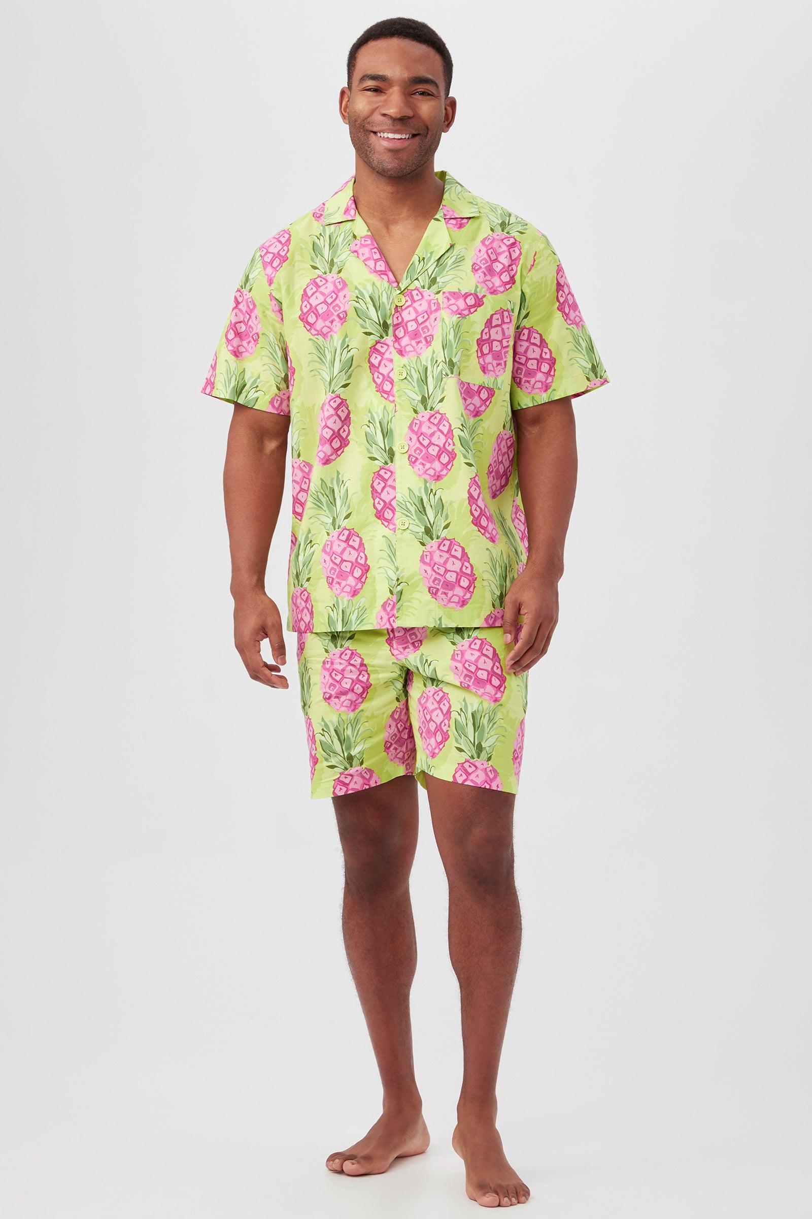A smiling man stands barefoot wearing the Kiwi Pineapple Mens Short Sleeve Boxer Short Poplin PJ Set in multi, featuring a bright green and pink pineapple print on a light background.